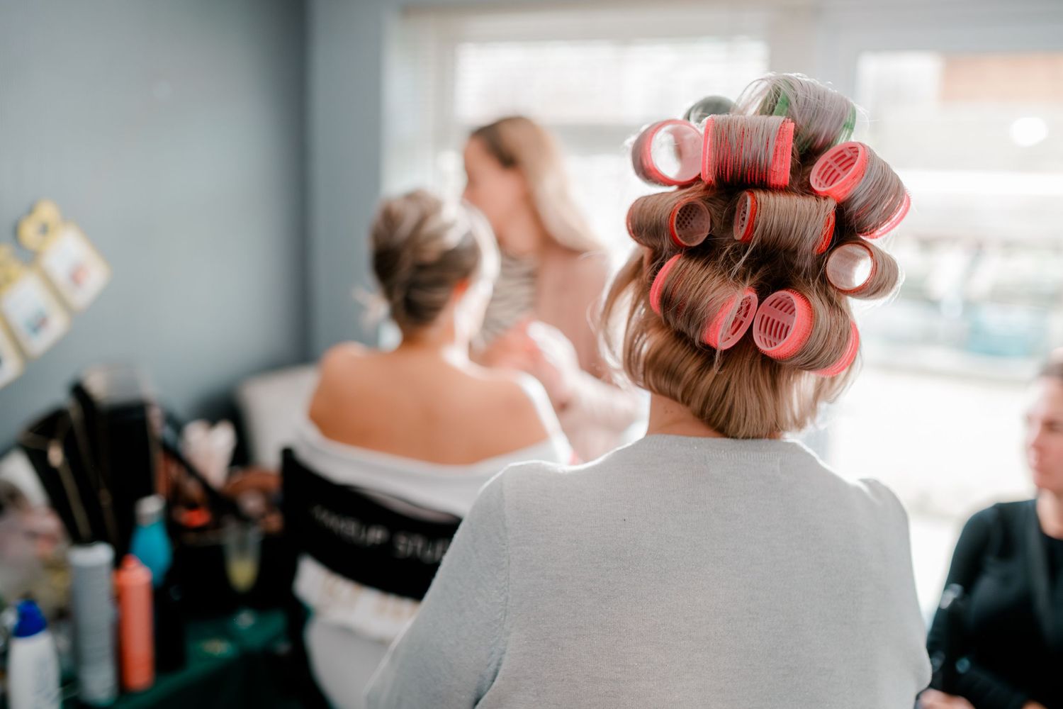 Back view of bridesmaid with pink hair rollers sitting in salon chair during wedding prep.