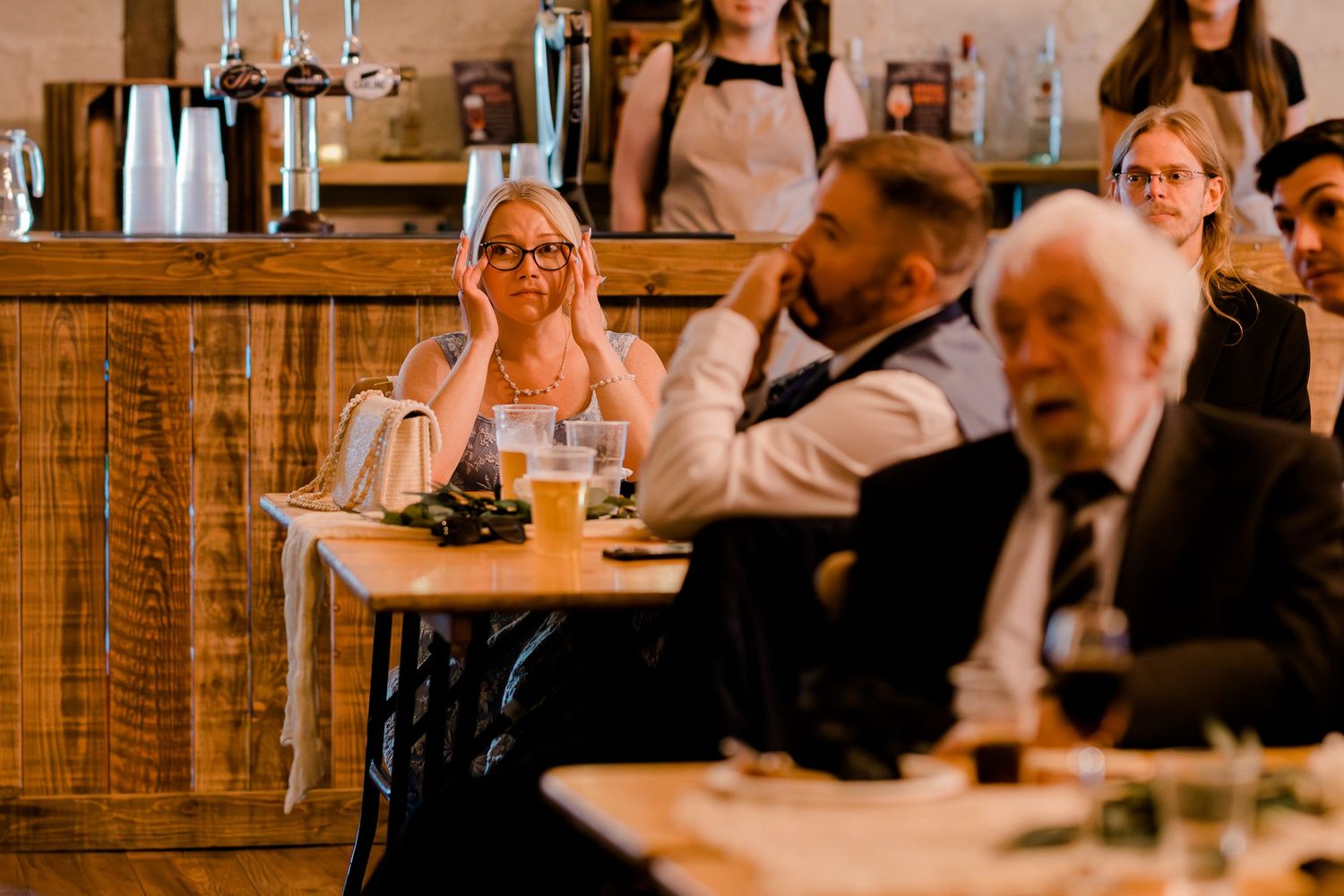 Wedding guests laughing and reacting emotionally during speeches at a rustic barn reception venue.