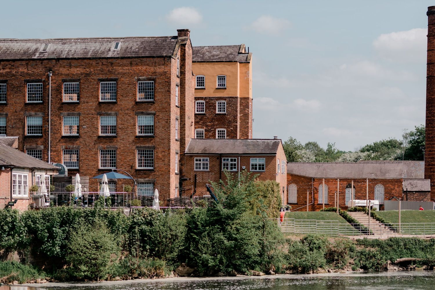Historic red brick mill building beside a river with lush greenery and outdoor seating area on a sunny day.