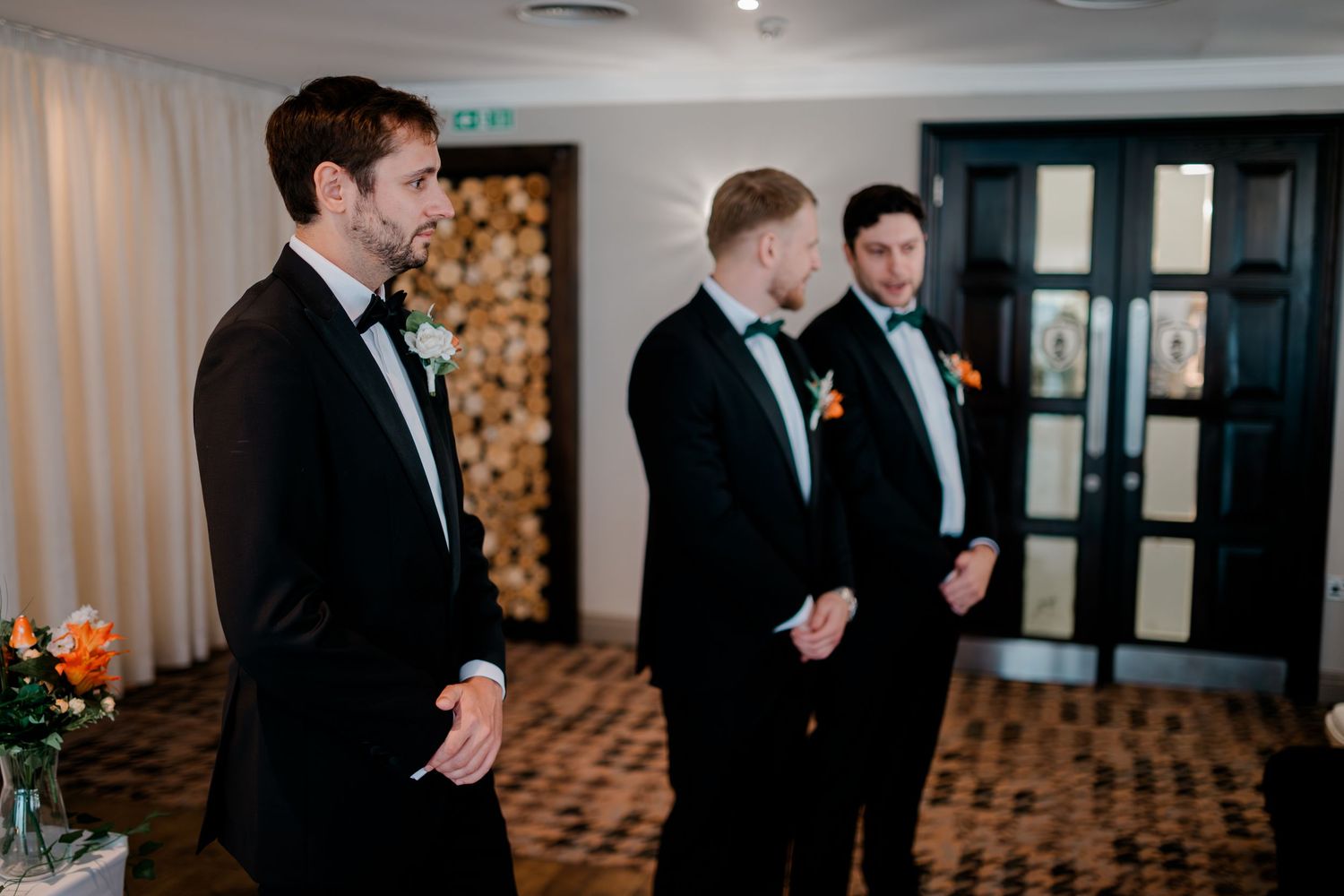 Three groomsmen in black tuxedos share a candid moment during a wedding ceremony in an elegant indoor venue.