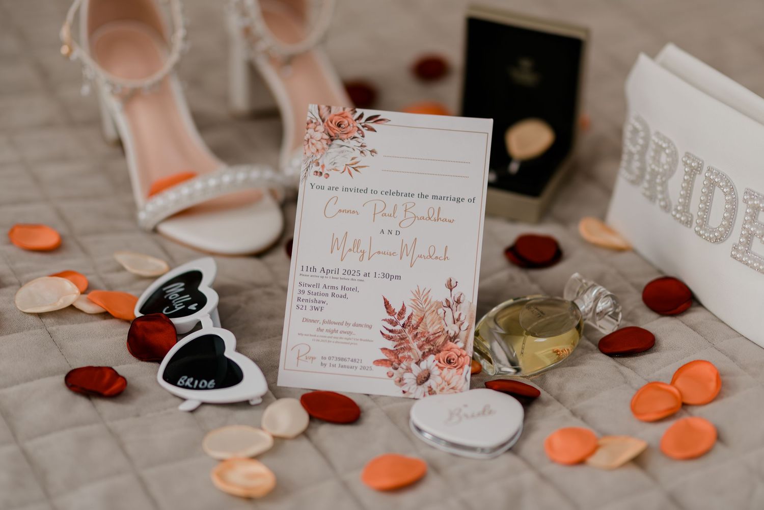 Wedding invitation with floral details surrounded by rose petals, sandals and decorative elements on a light textile surface.