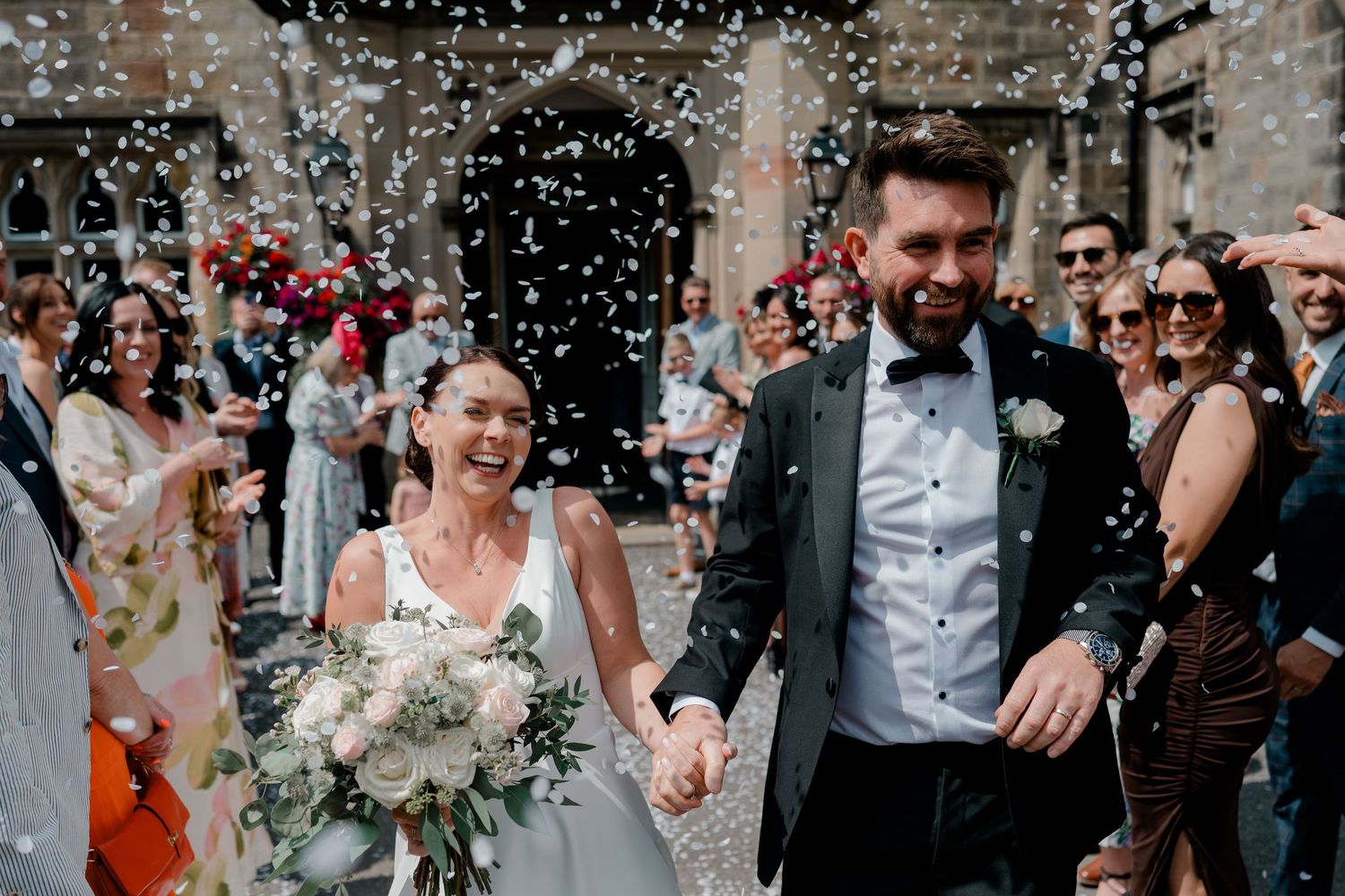 Smiling bride with bouquet and groom in bow tie walk hand in hand through confetti outside a stone church as wedding guests cheer.