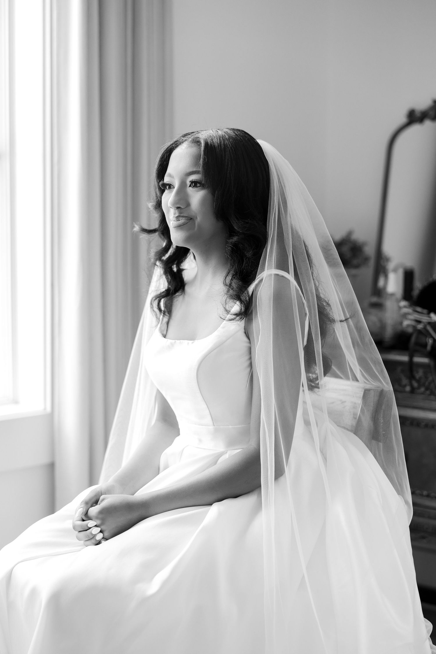 Black and white portrait of a bride in a white gown and veil, seated gracefully by a window on her wedding day.