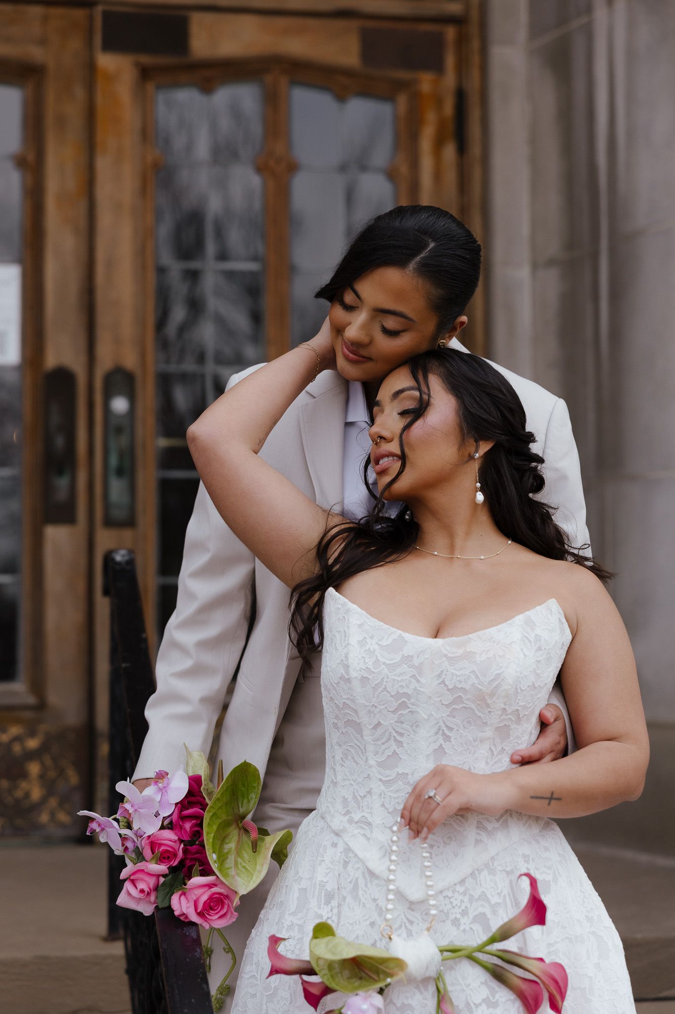 A bride in a lace wedding dress poses with pink roses against a stone architectural backdrop.
