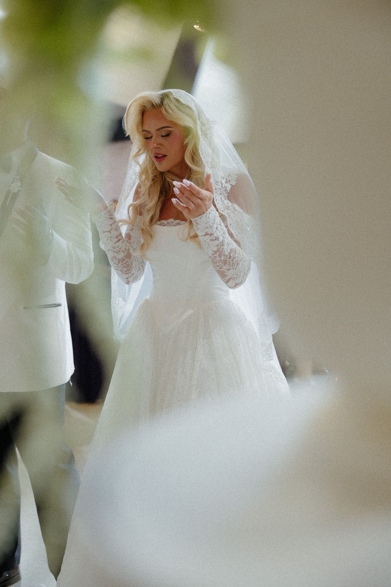 Elegant blonde bride in white lace wedding dress and veil, hands clasped, standing in soft natural light.