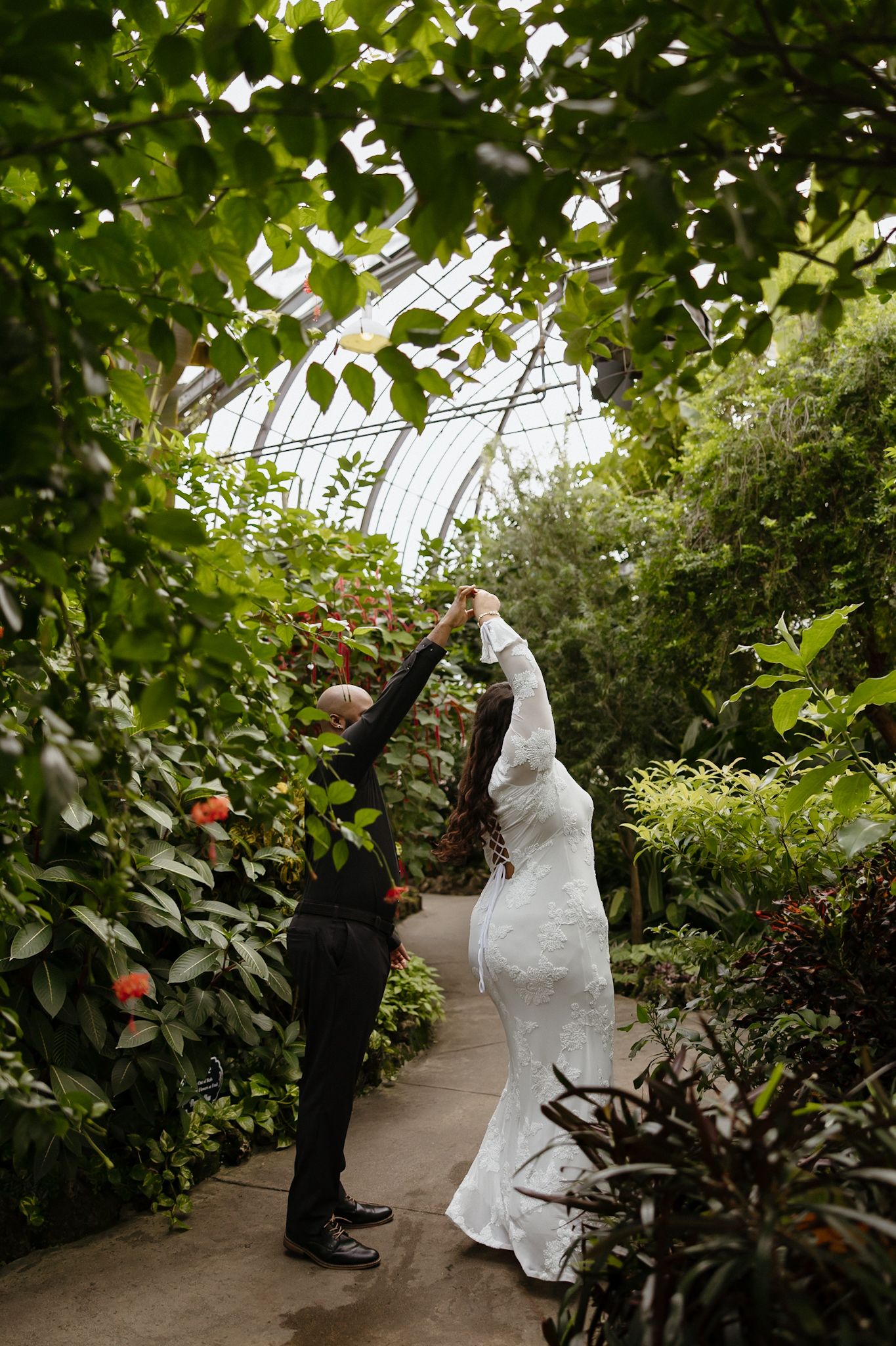 A couple dances playfully in a greenhouse filled with lush greenery and natural light.