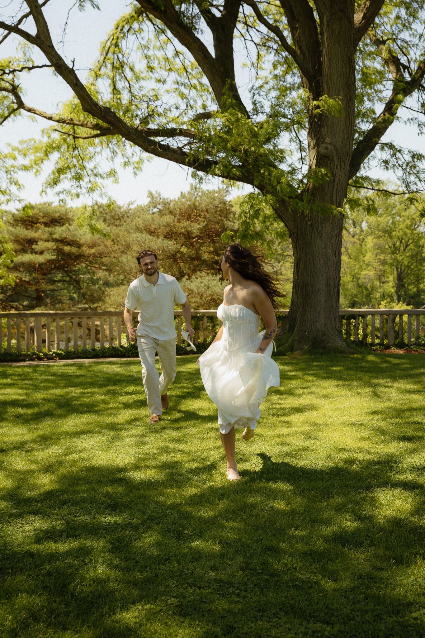 A joyful couple in white attire running barefoot on lush green grass near a large tree on a sunny day.