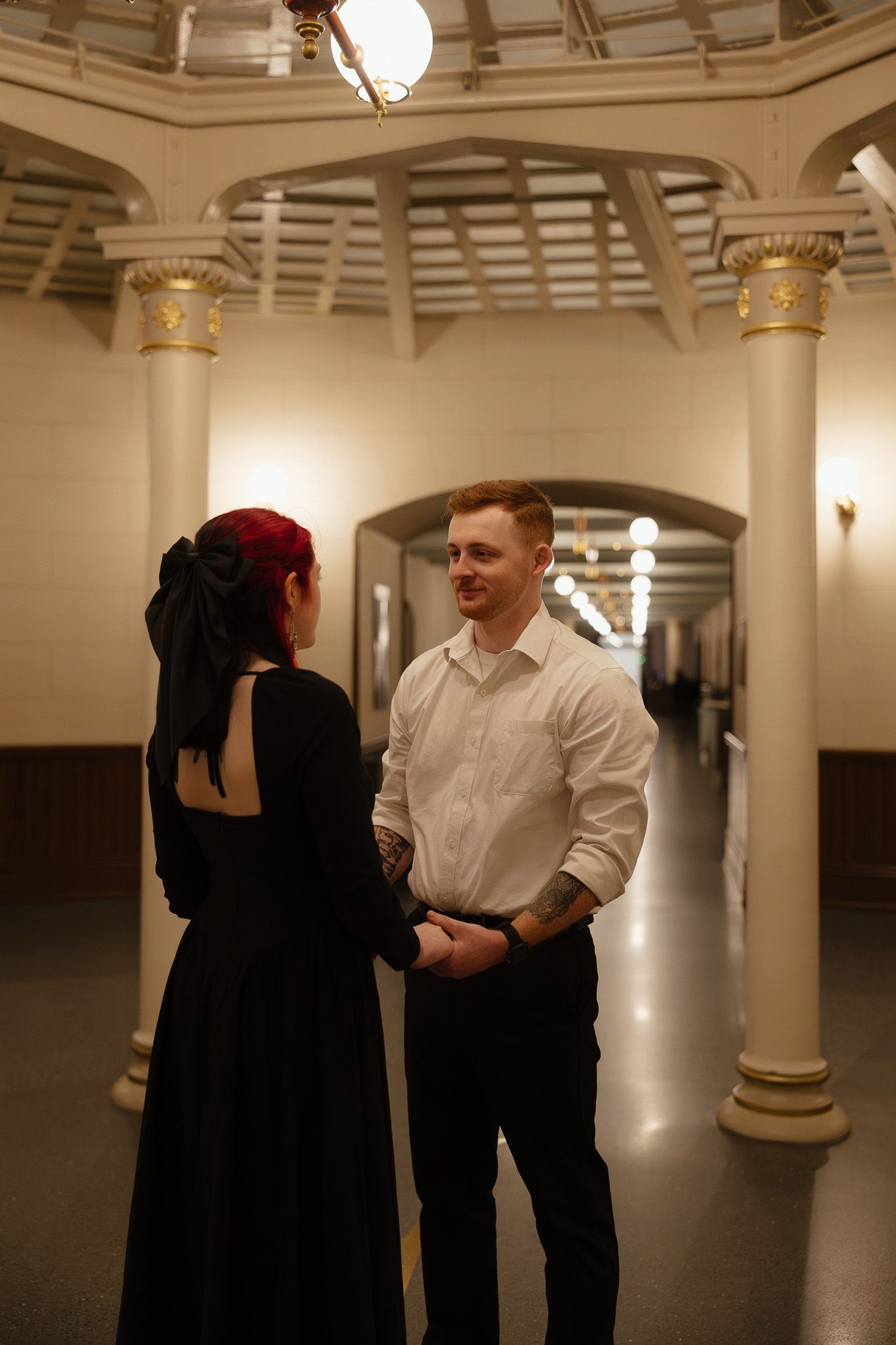 Elegant couple holding hands in an ornate hallway with columns, woman in black backless dress.