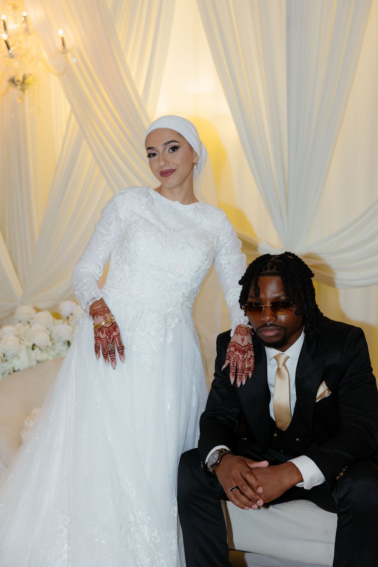 Bride in white hijab gown and groom in black suit pose together at elegant wedding ceremony.