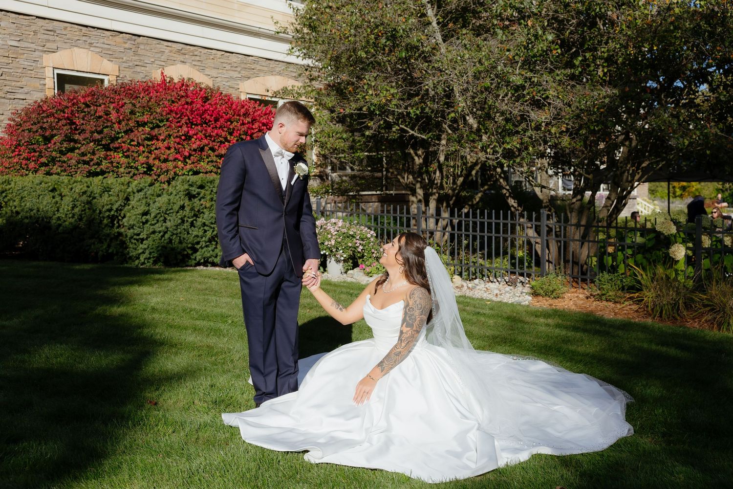 Bride in white gown sits on green lawn holding groom's hand in navy suit, with red flowers and trees in background.