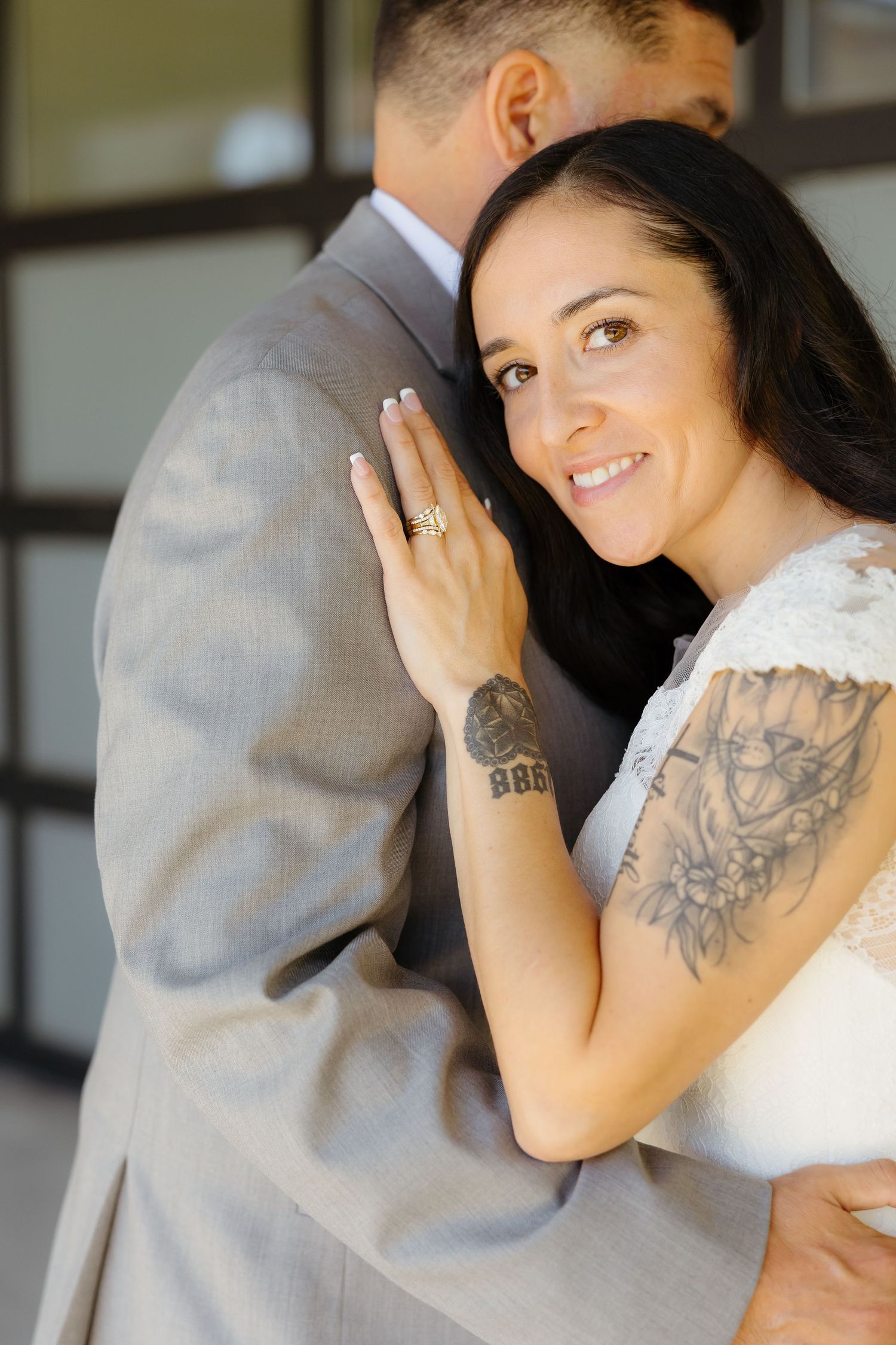 Smiling bride with tattoos hugs groom in gray suit, showing off engagement ring on her hand at wedding.