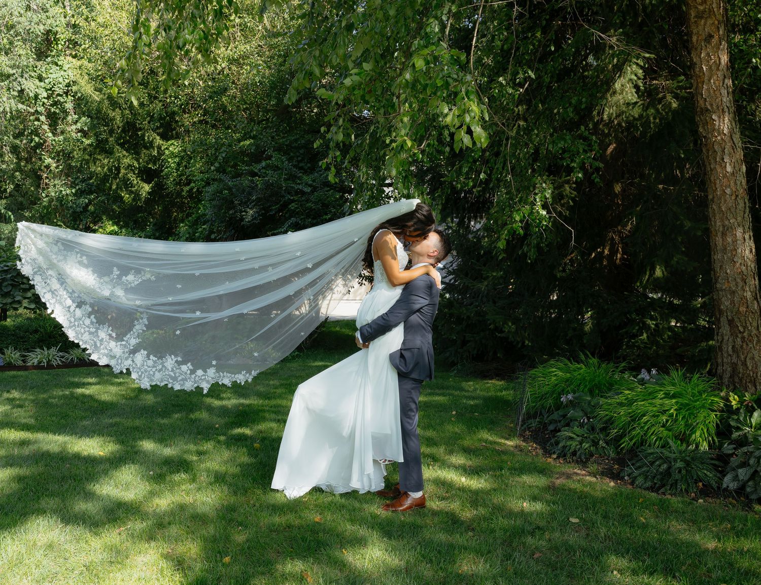 Groom lifting bride in white gown and flowing veil during outdoor wedding ceremony in lush green garden.