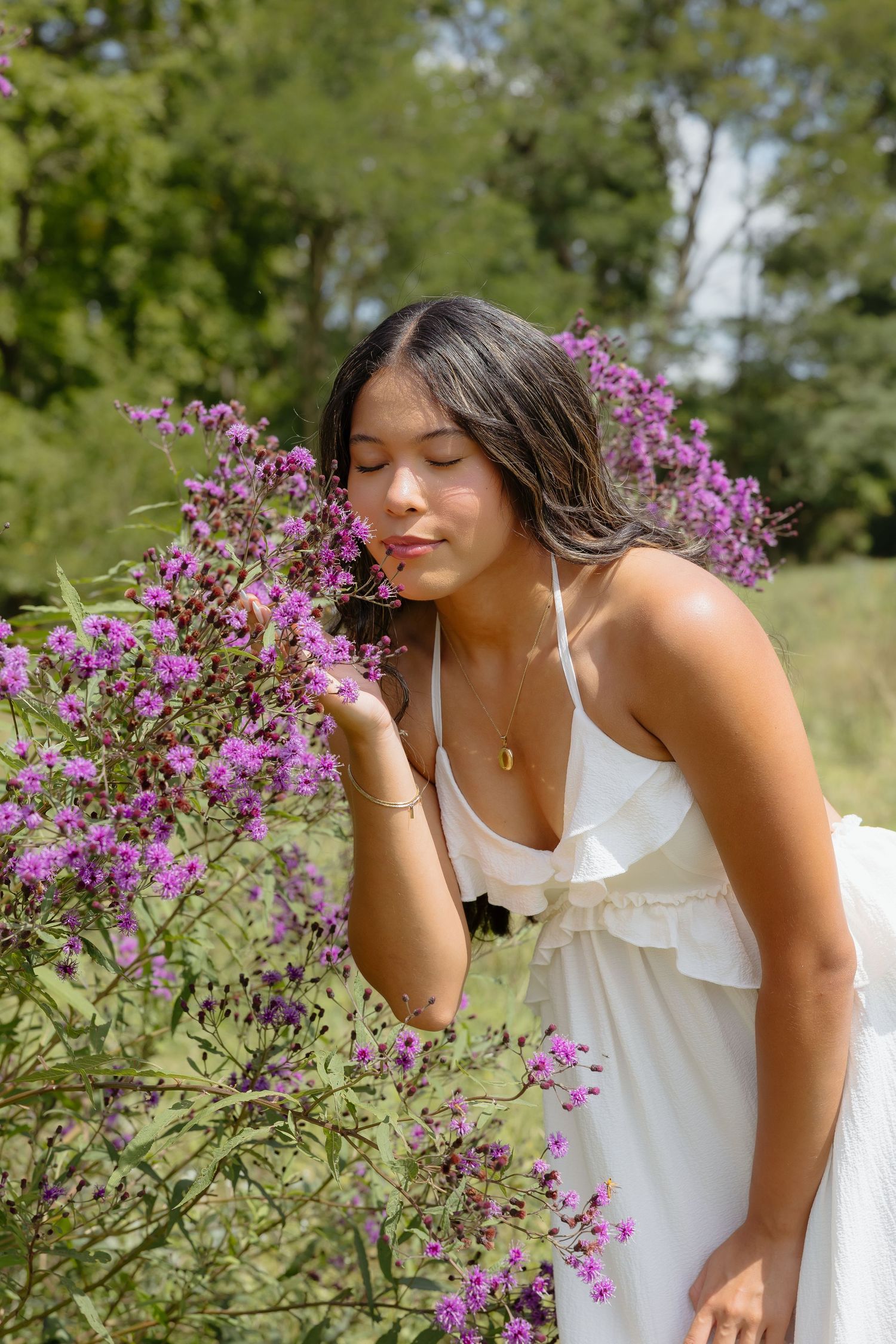 Young woman in white dress smelling purple wildflowers outdoors with eyes closed, surrounded by lush green trees.