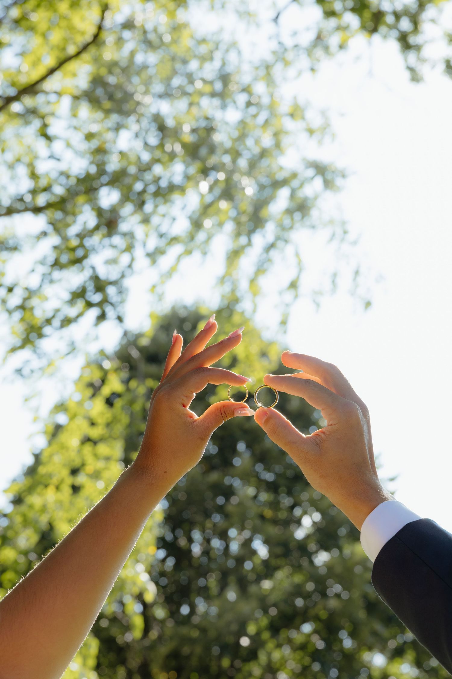 Couple exchanging gold wedding rings outdoors, hands reaching up toward sunlit green trees during marriage ceremony.