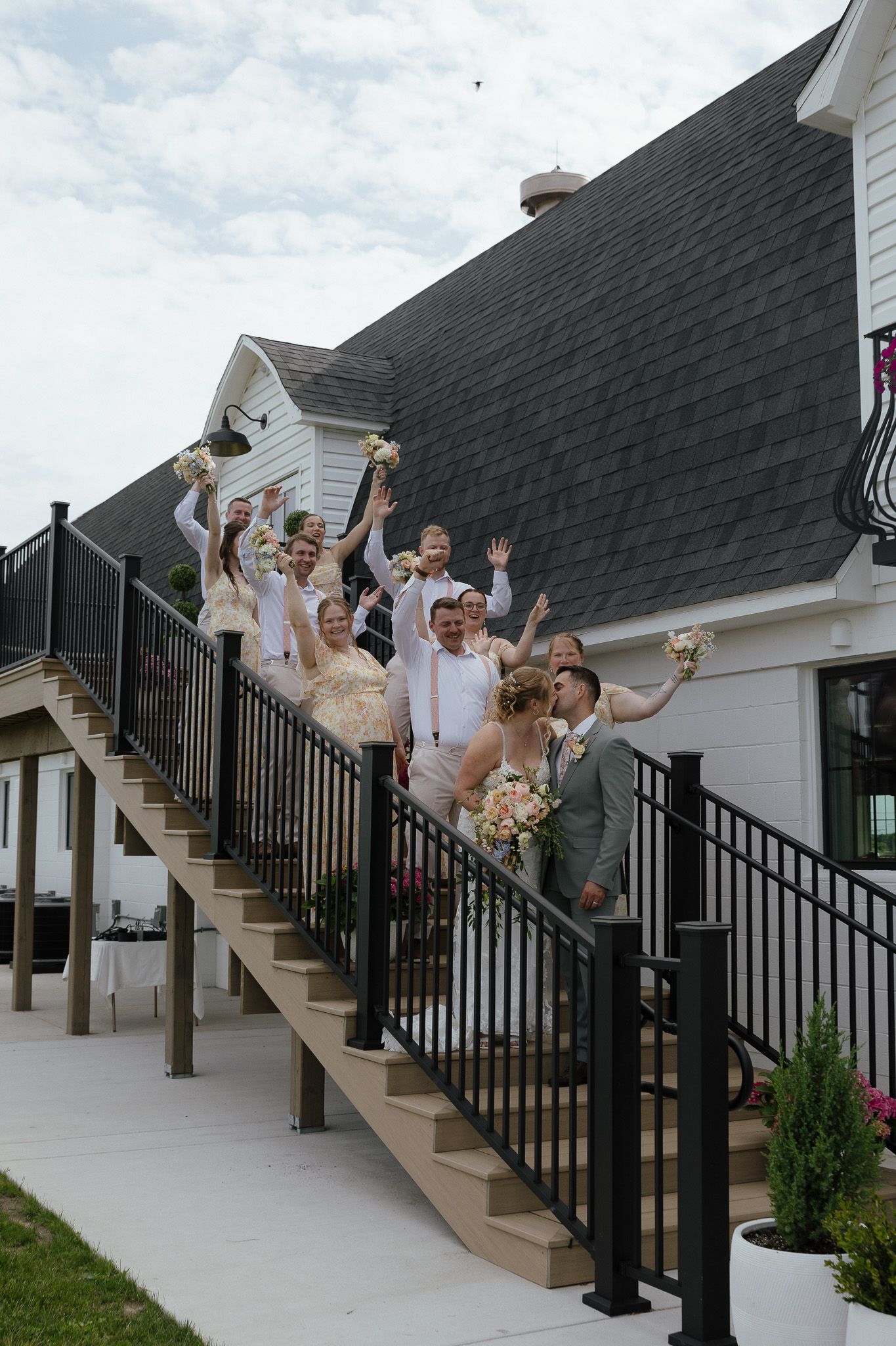 Wedding party celebrating on outdoor staircase of white venue, waving bouquets under cloudy sky.