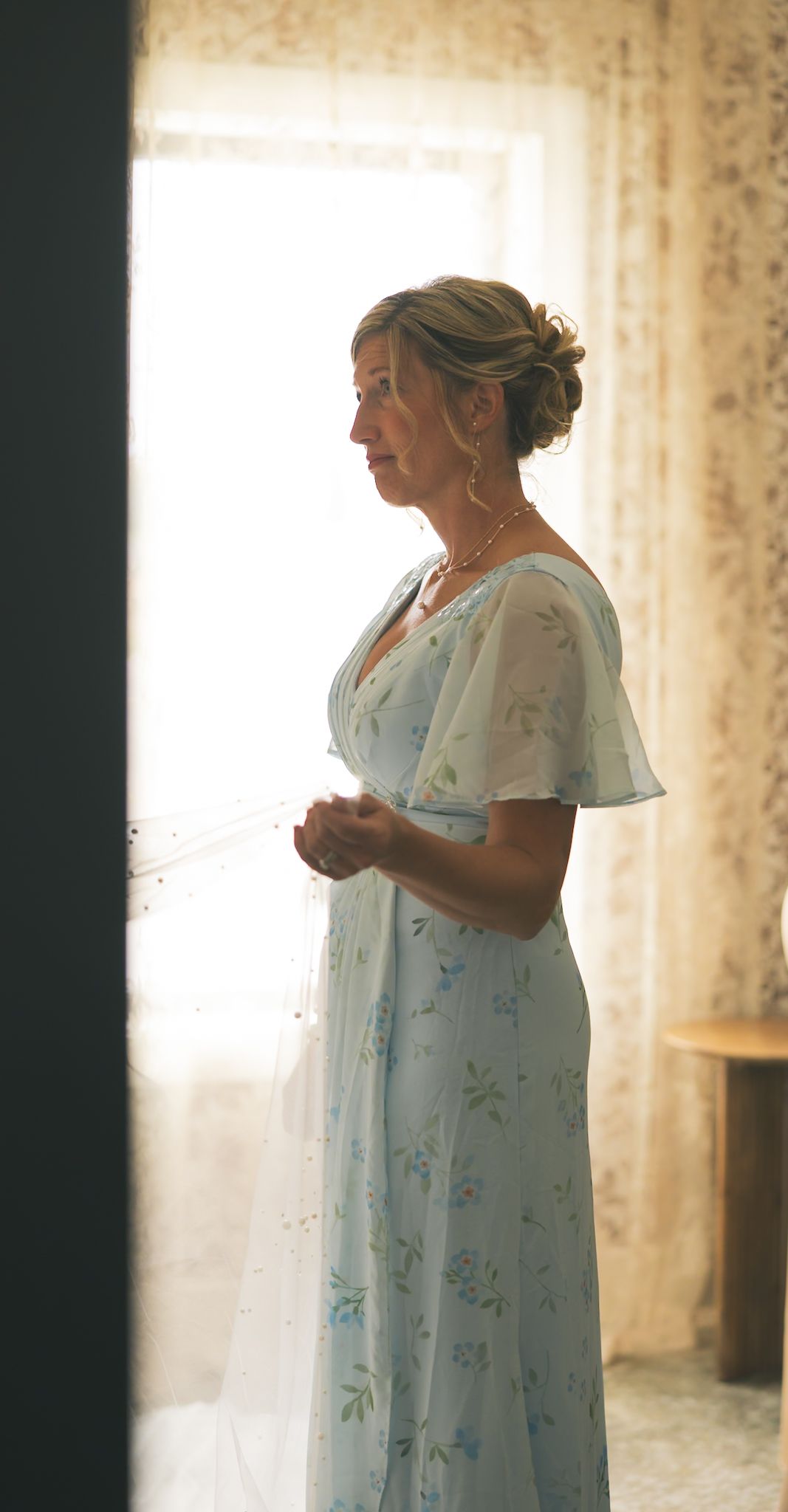 Someone in a floral white nightgown stands in front of sheer curtains in a sunlit room.