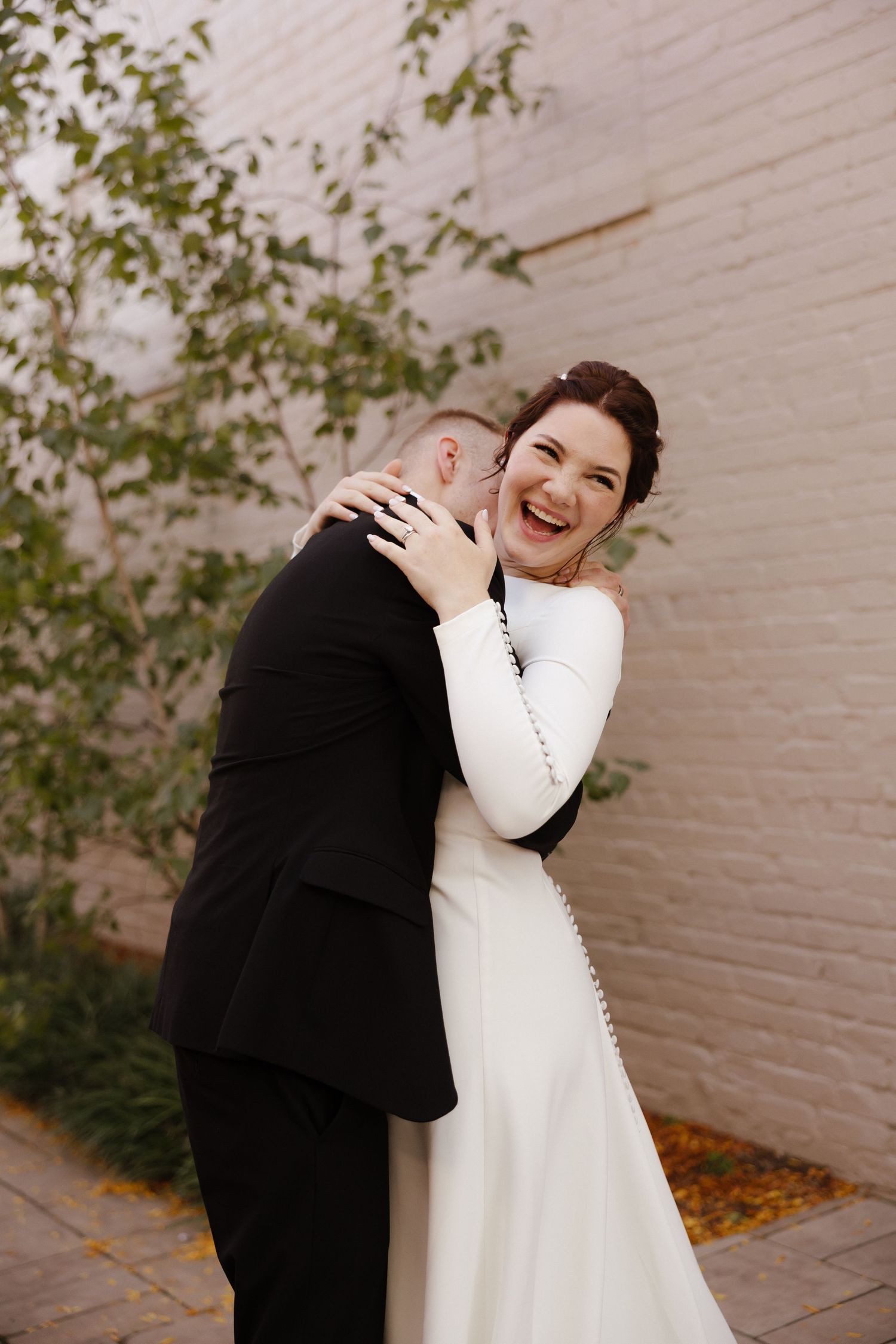 Joyful wedding couple embraces against white brick wall with autumn leaves.