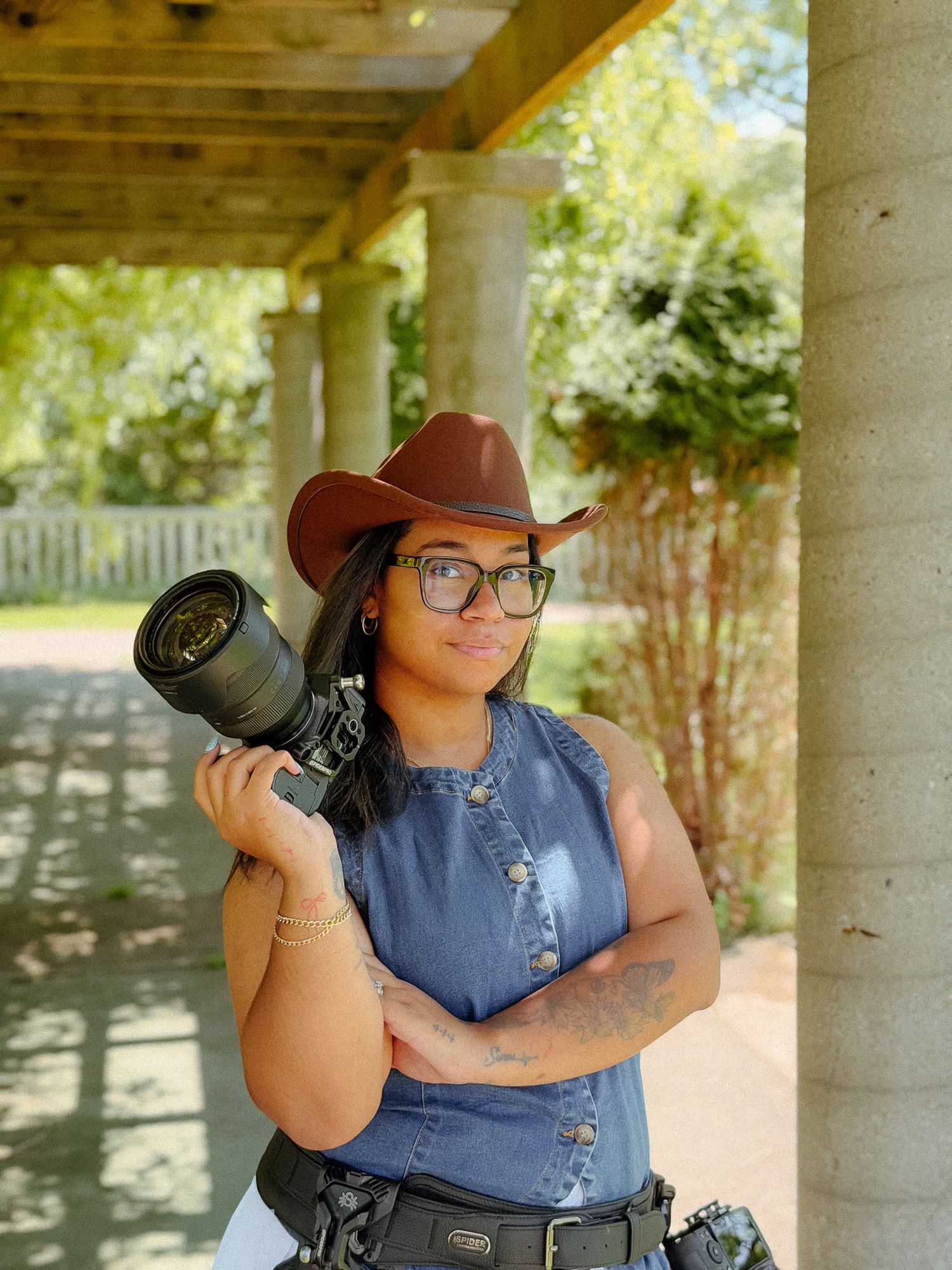 A young woman in a brown cowboy hat and denim vest holds a professional camera outdoors near stone columns.