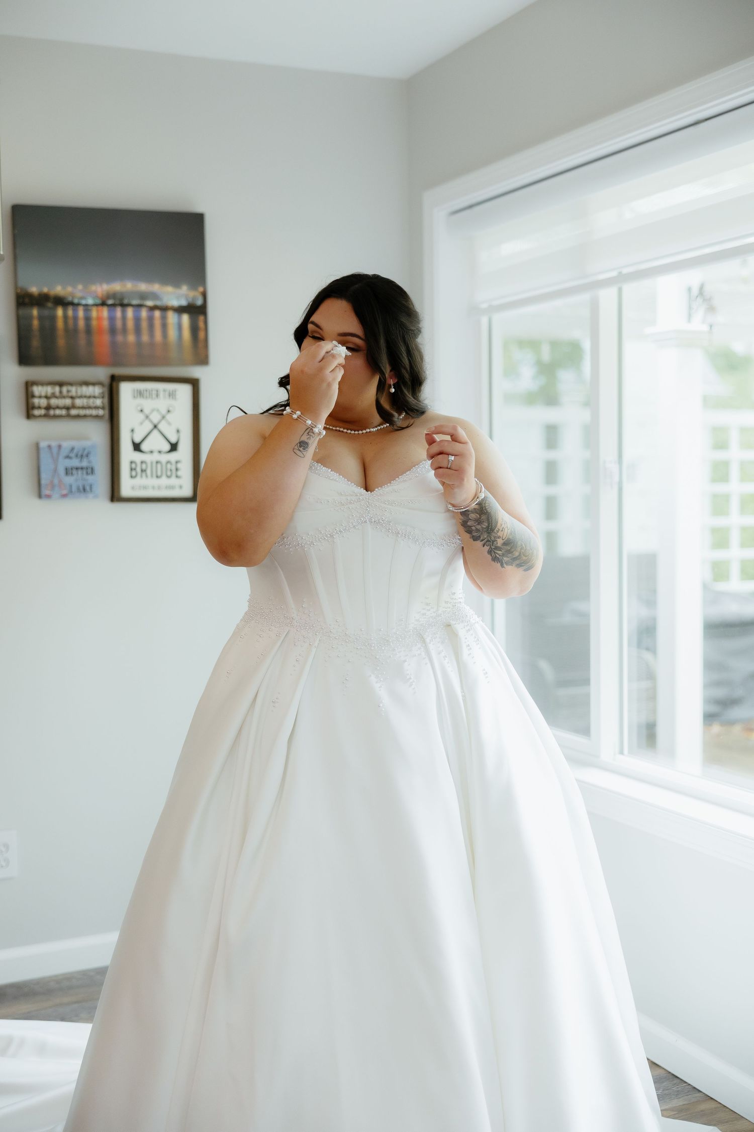 Emotional bride in a white strapless ball gown wiping tears, with tattoos visible, standing near a bright window.