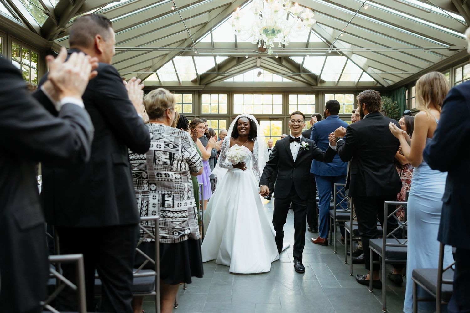 Bride and groom walk down the aisle in a sunlit greenhouse wedding venue, surrounded by applauding guests.