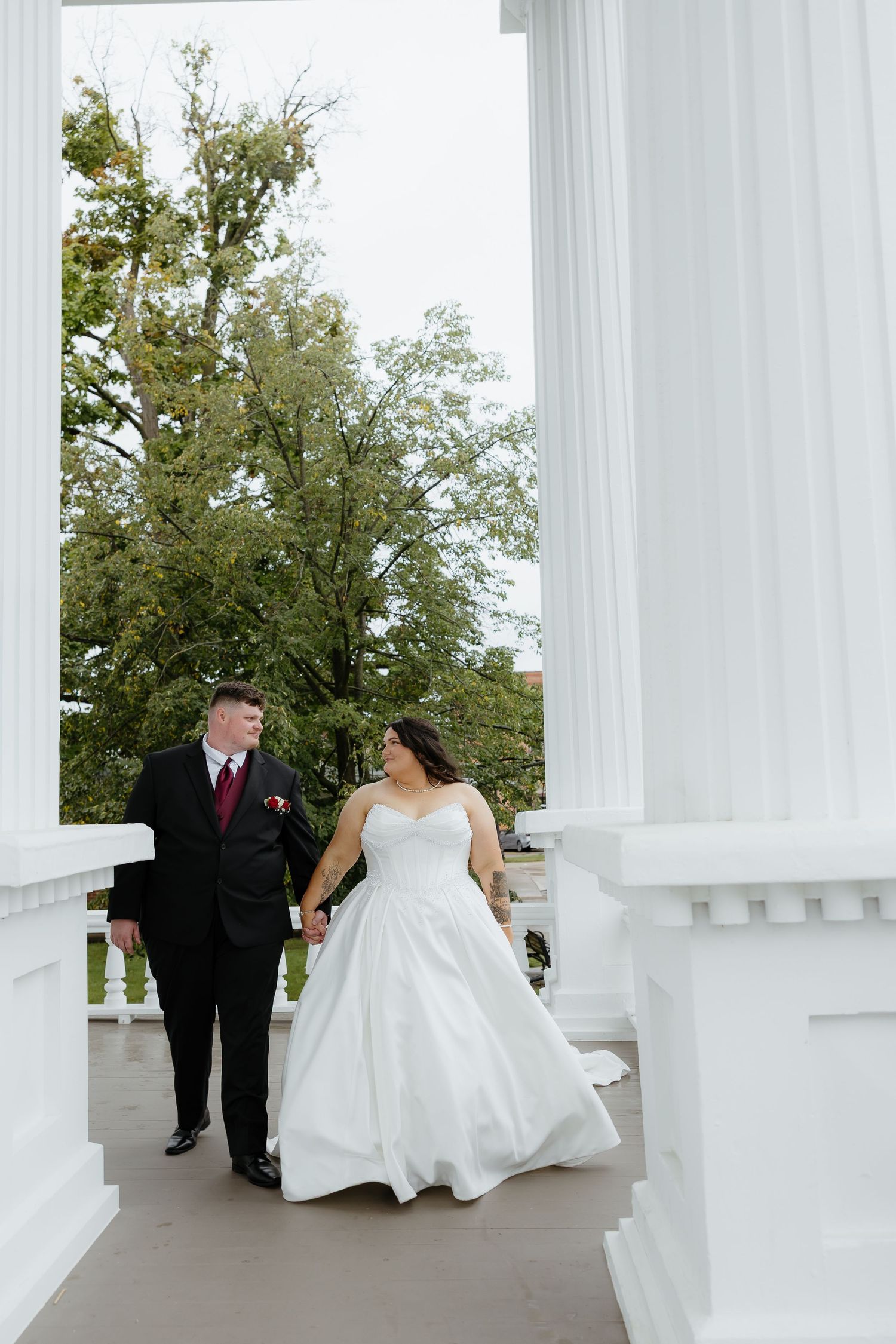 Bride and groom walk hand-in-hand between white columns on a porch, smiling at each other in wedding attire.