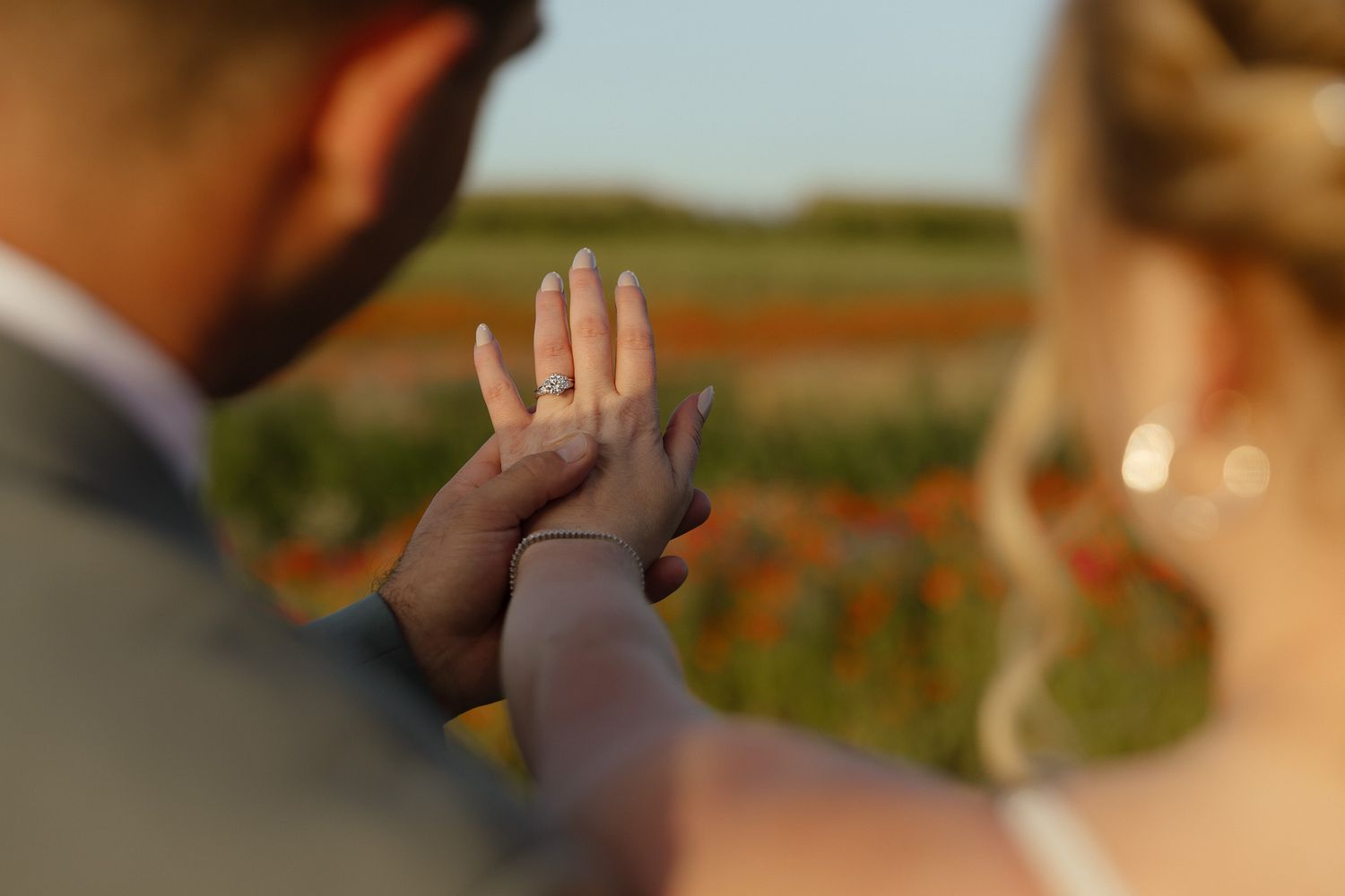 Bride and groom holding hands displaying wedding ring in golden sunlight with colorful flower field in background.