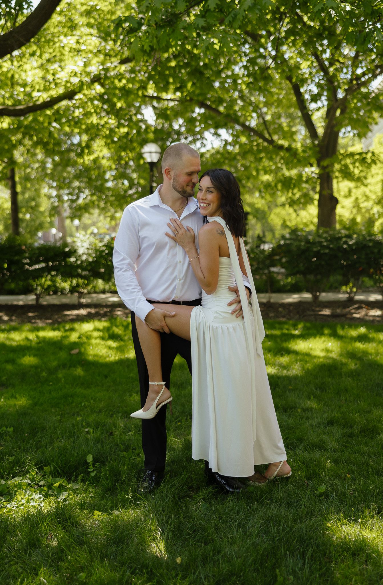 Couple in wedding attire embracing in a lush green park, bride in white dress smiling joyfully.