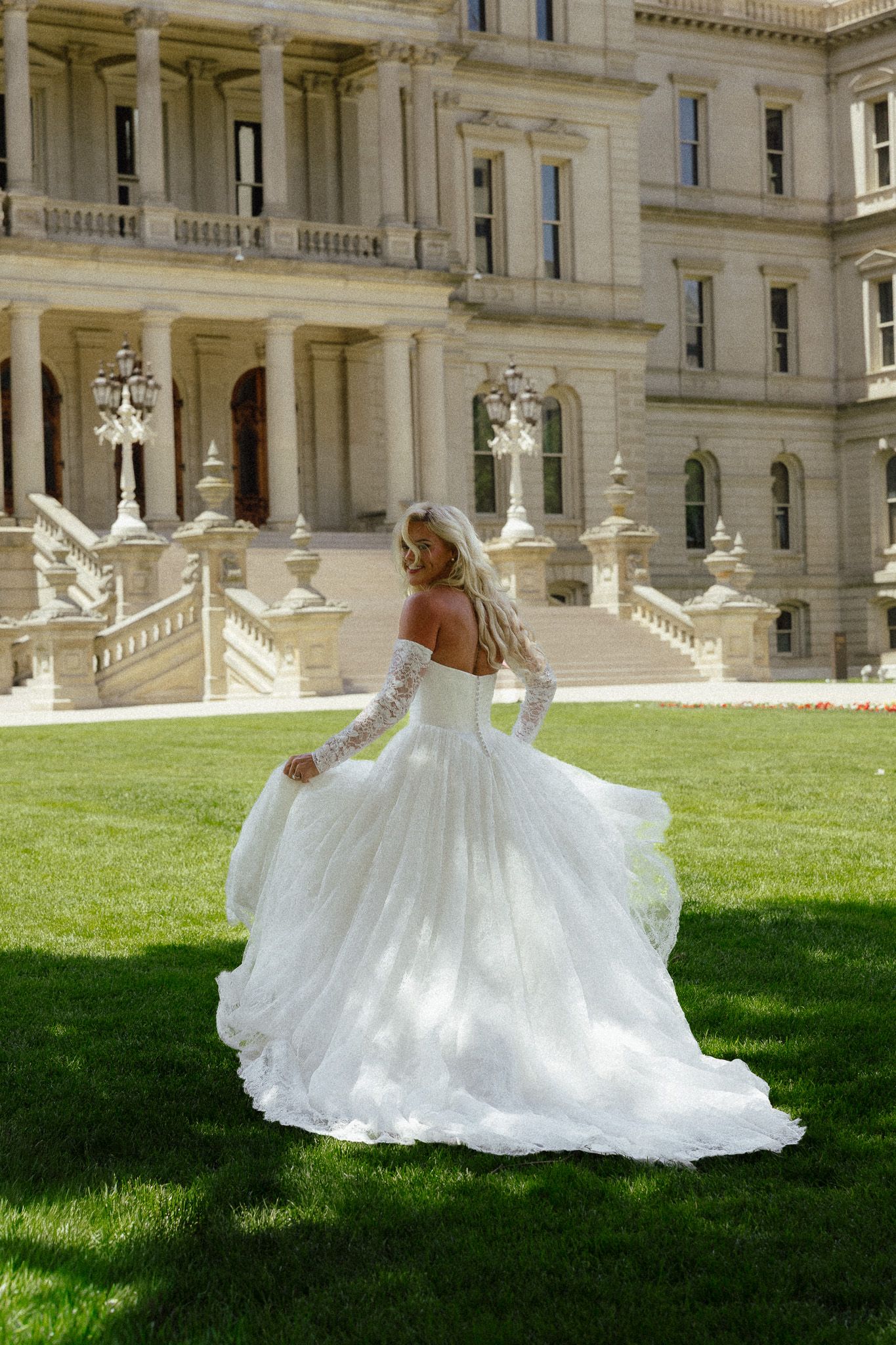 Bride in elegant white off-shoulder ball gown with lace sleeves twirling on green lawn before grand stone building.