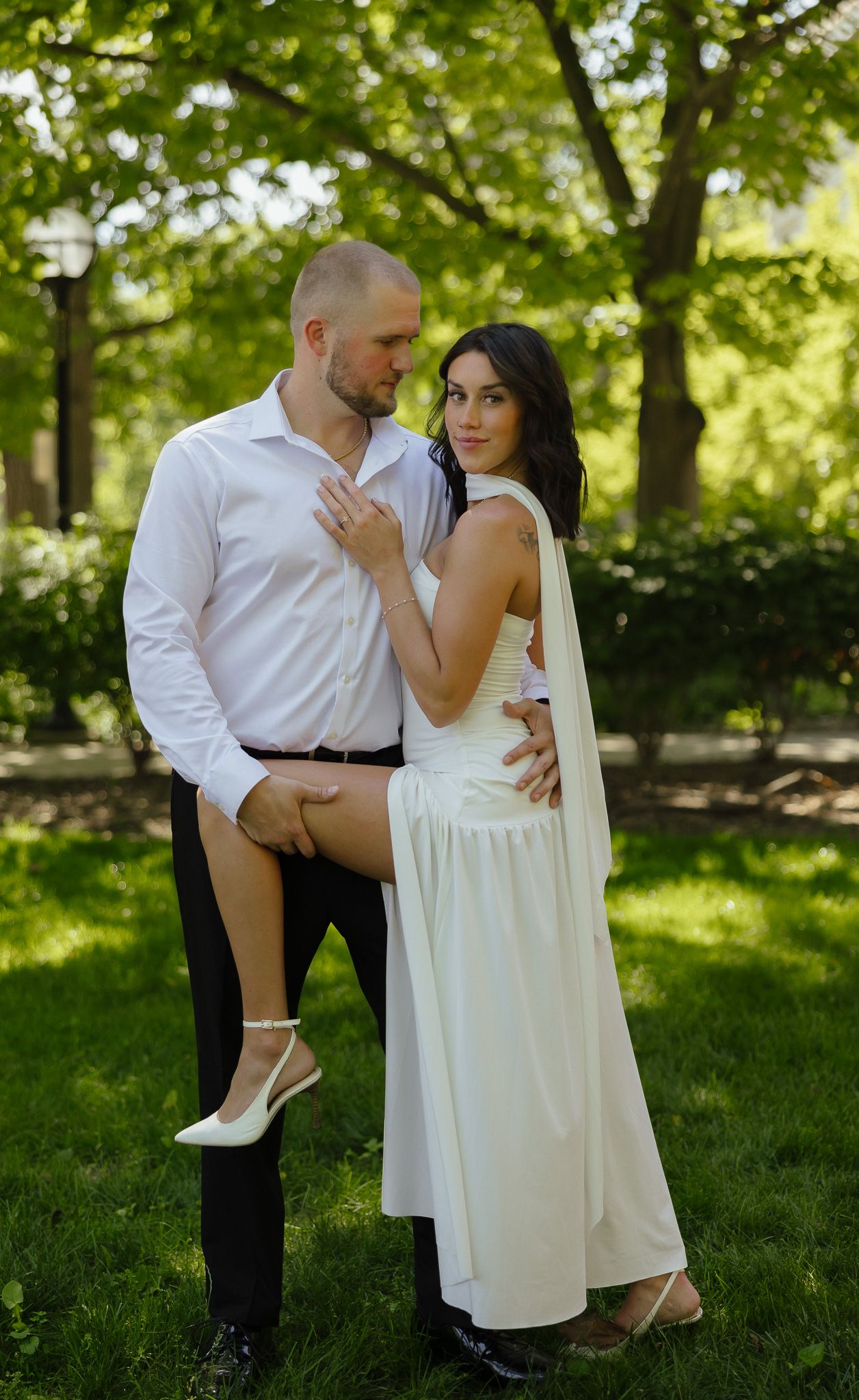 Couple in wedding attire posing outdoors in a green park, bride in white dress, groom in white shirt and black pants.