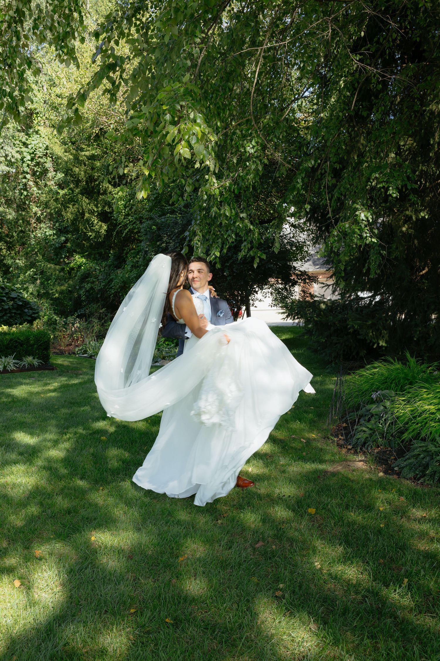 Groom spinning bride in white gown and veil in lush green garden on sunny wedding day.