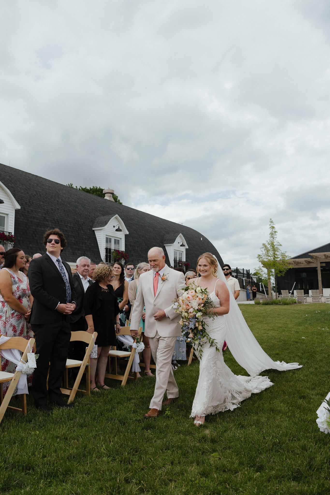 Bride in lace gown walks down outdoor aisle with escort, carrying a cascading bouquet at a garden wedding ceremony.
