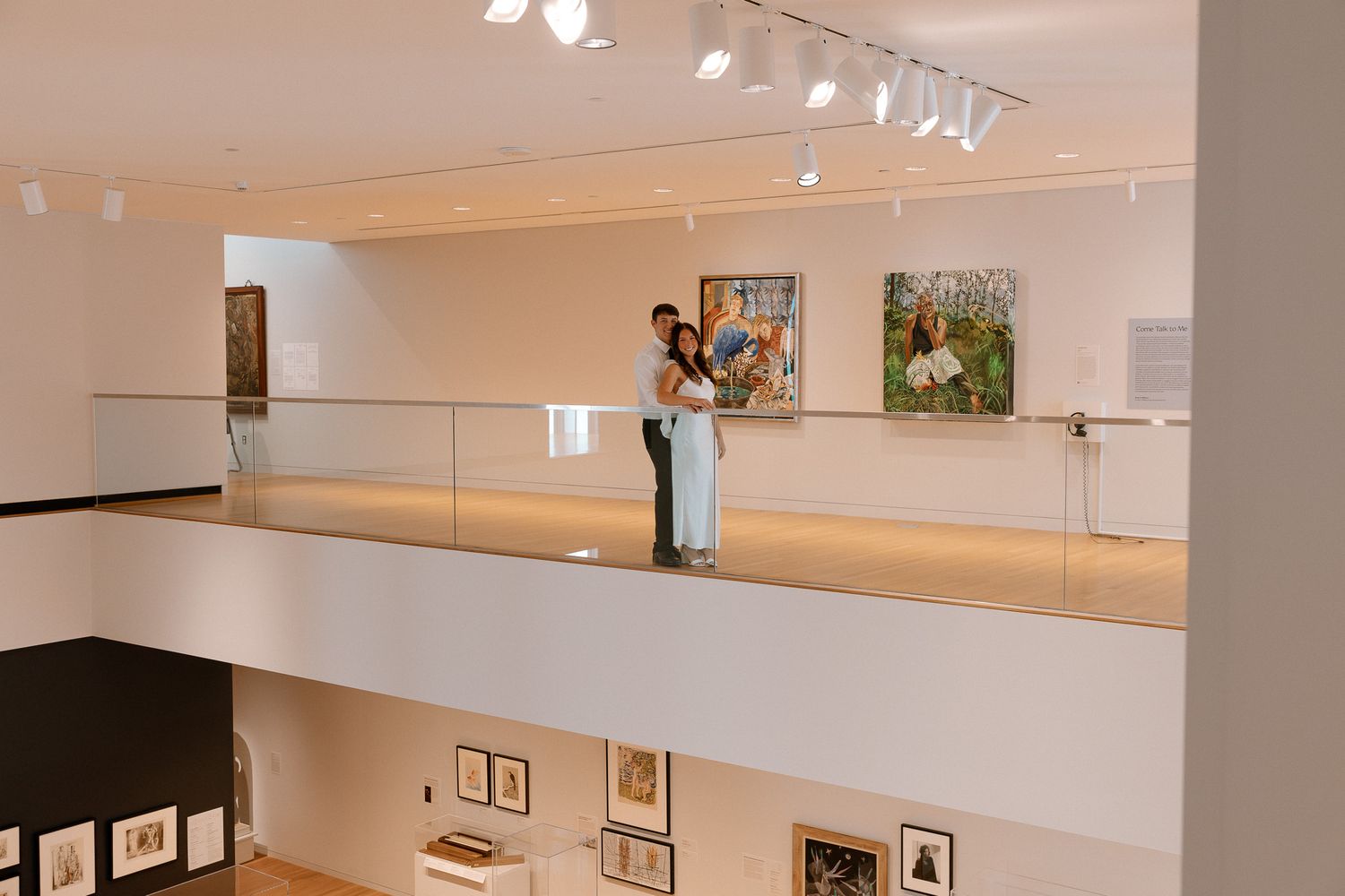 Couple embracing on a modern art museum balcony overlooking a gallery with framed artwork on white walls.