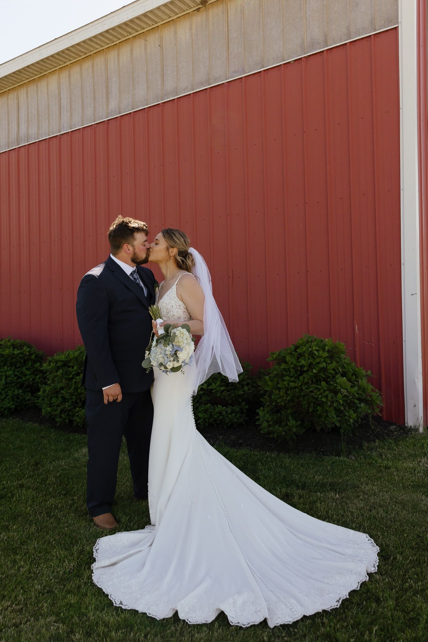 Bride and groom kissing in front of a red barn, bride wearing a white gown with veil holding a bouquet.