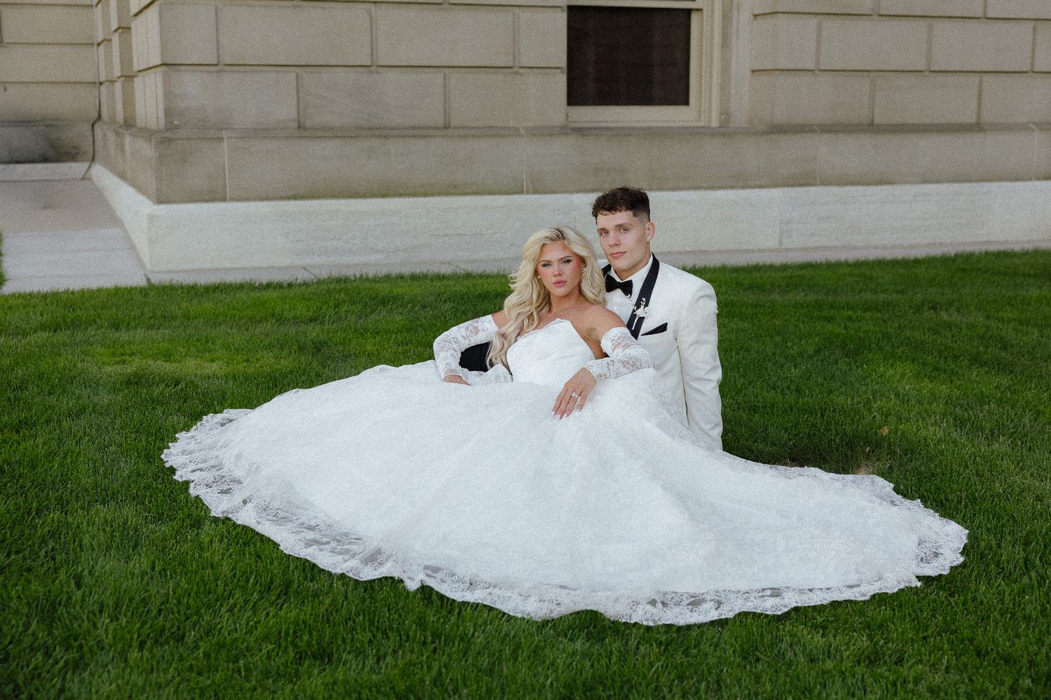 Bride in white ball gown and groom in white tuxedo sitting on green grass outside a stone building.