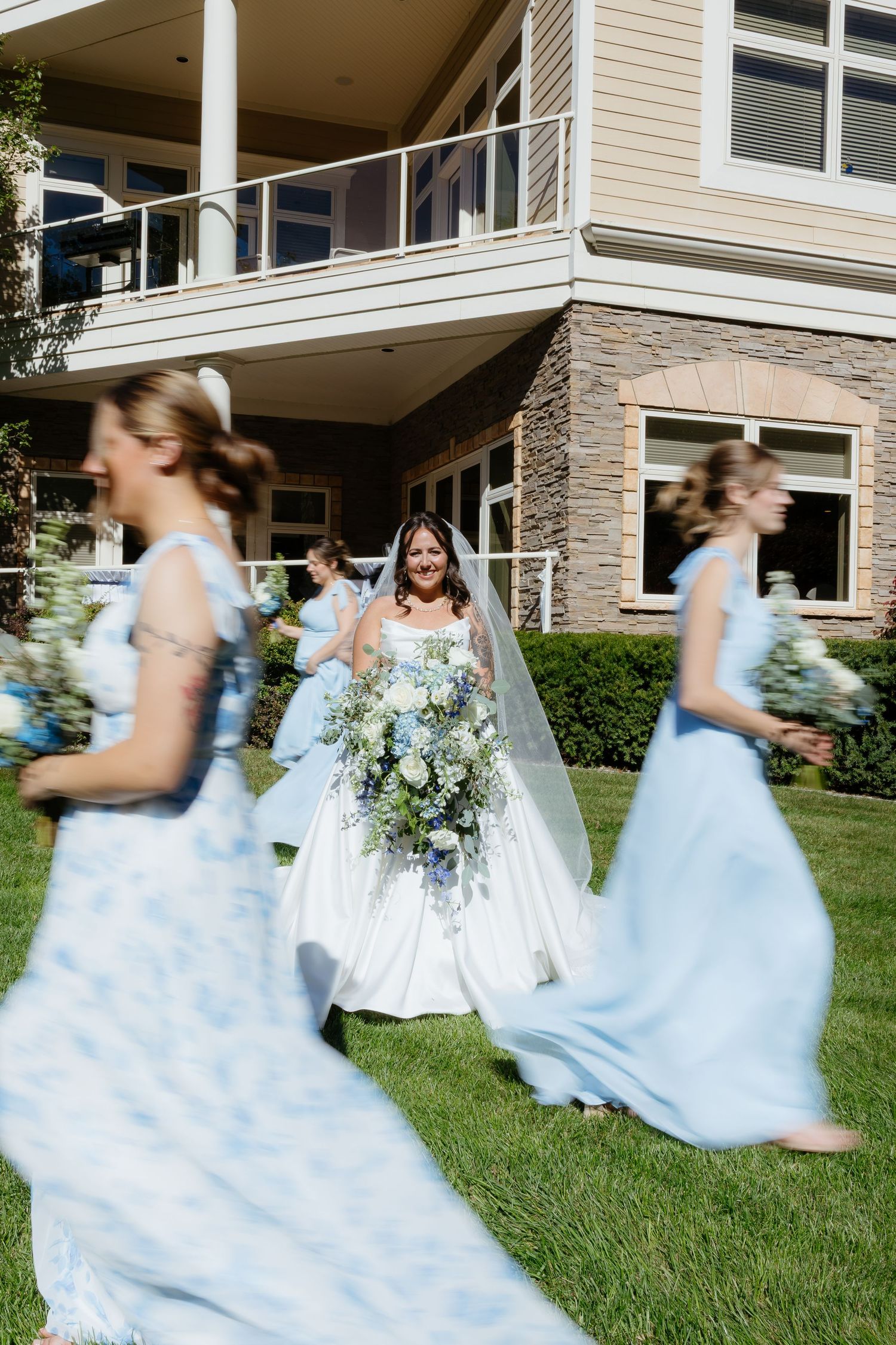 Bride in white gown holds blue floral bouquet surrounded by bridesmaids in light blue dresses on green lawn.