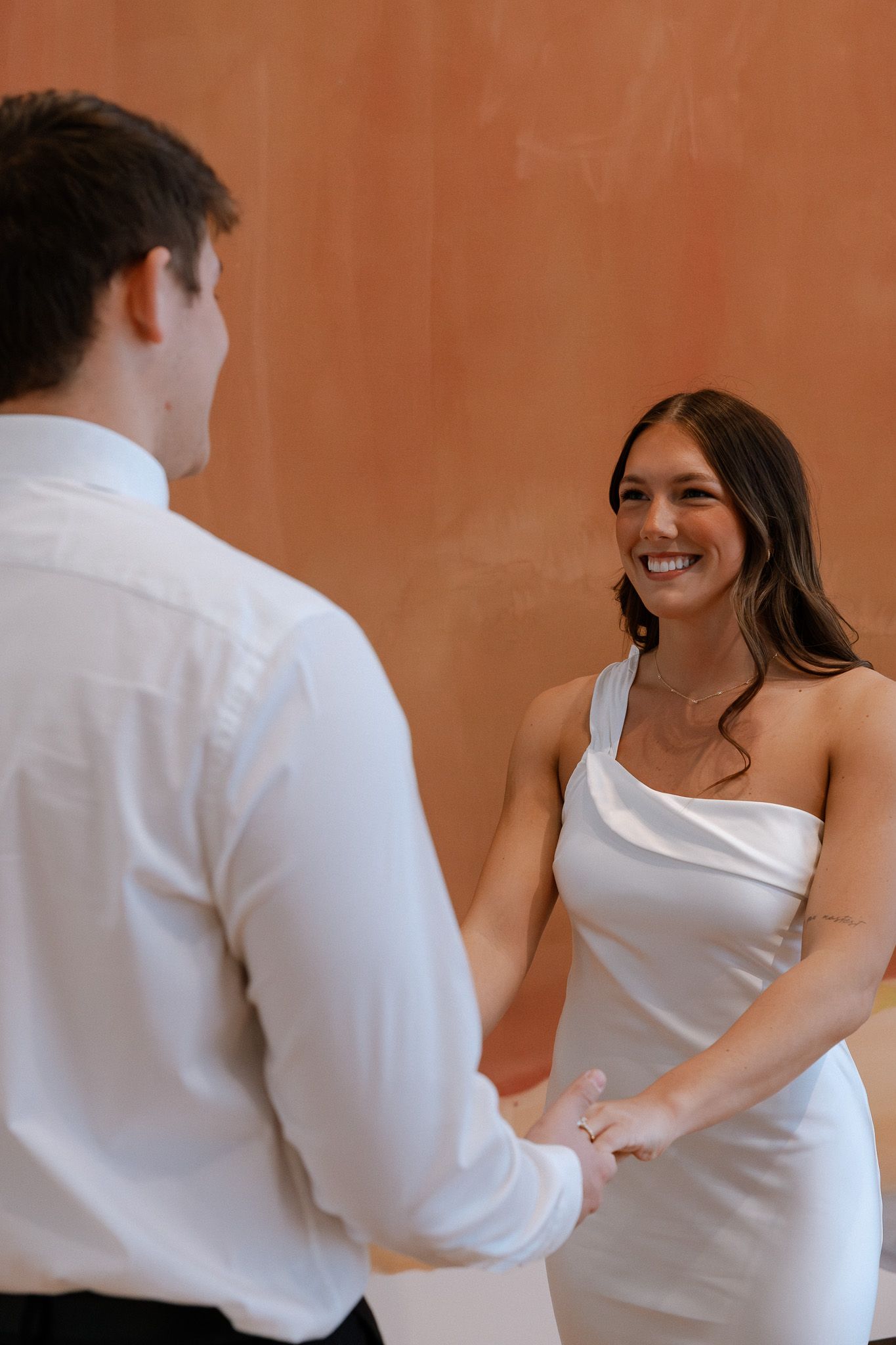 A smiling bride in a white dress holds hands with a groom in a white shirt against a warm terracotta wall.