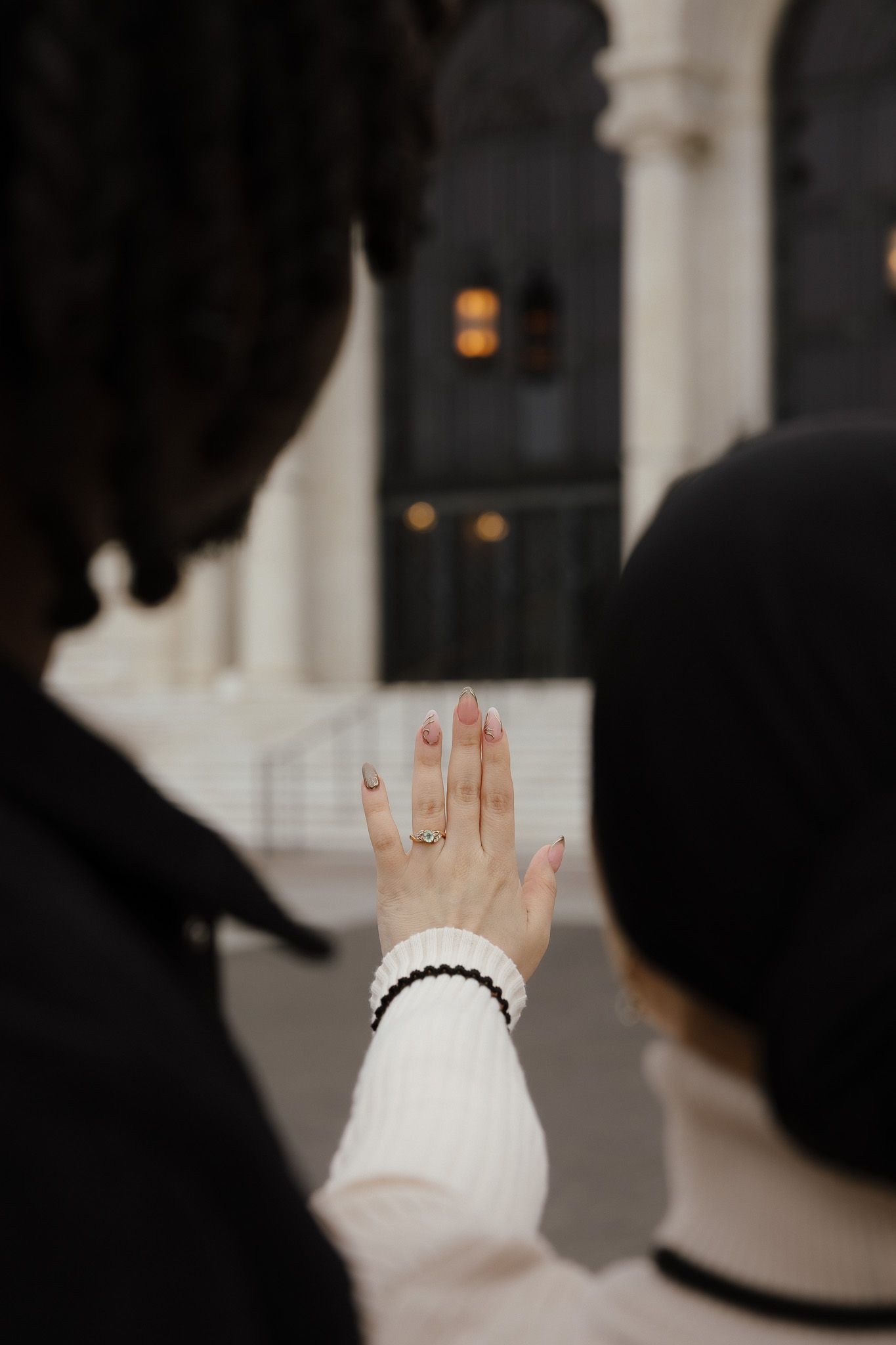 A woman displays her engagement ring while standing outdoors near an elegant building entrance.