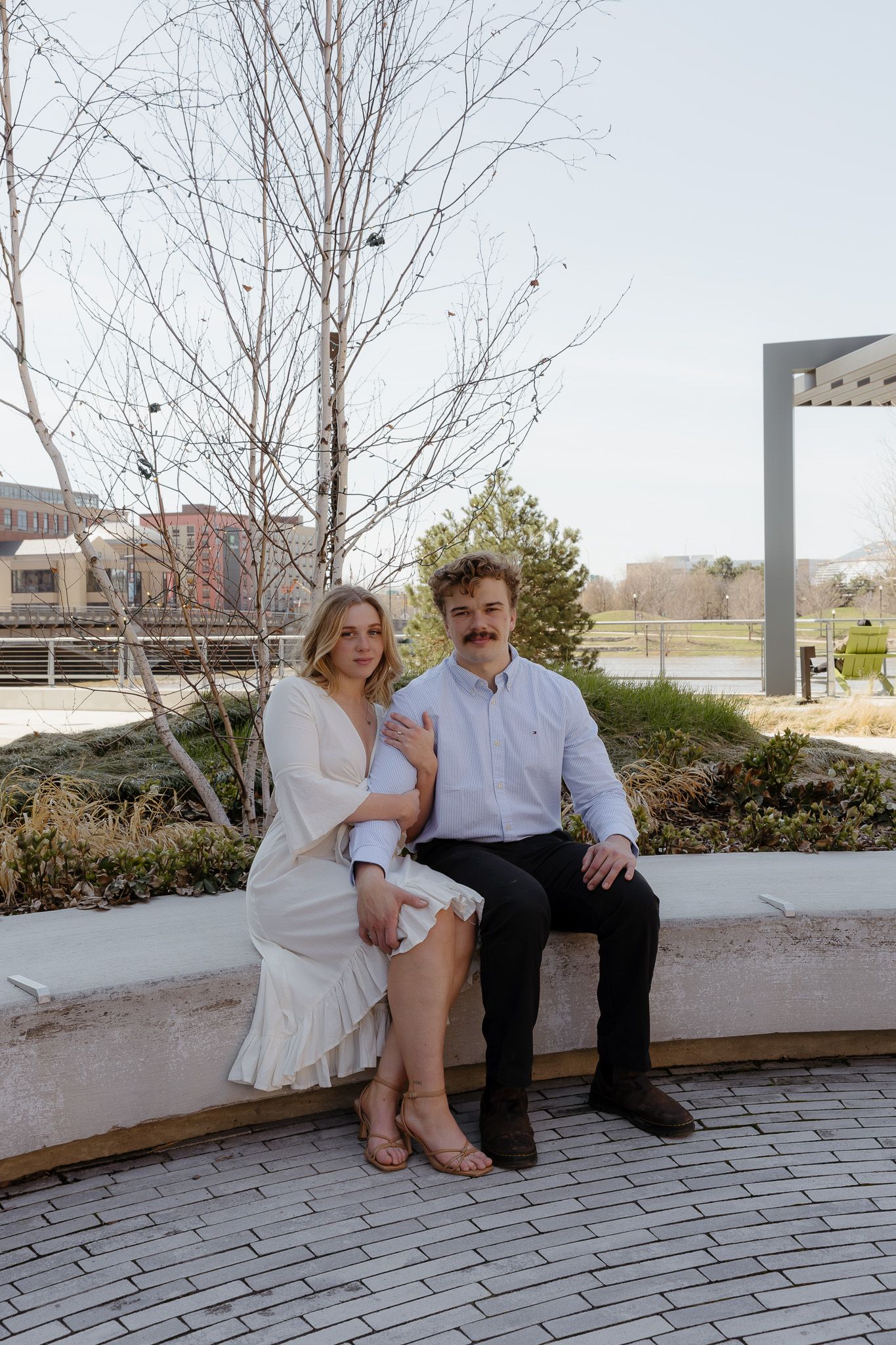 A couple sits together on a stone bench in an outdoor park setting, woman in white dress and man in blue shirt.