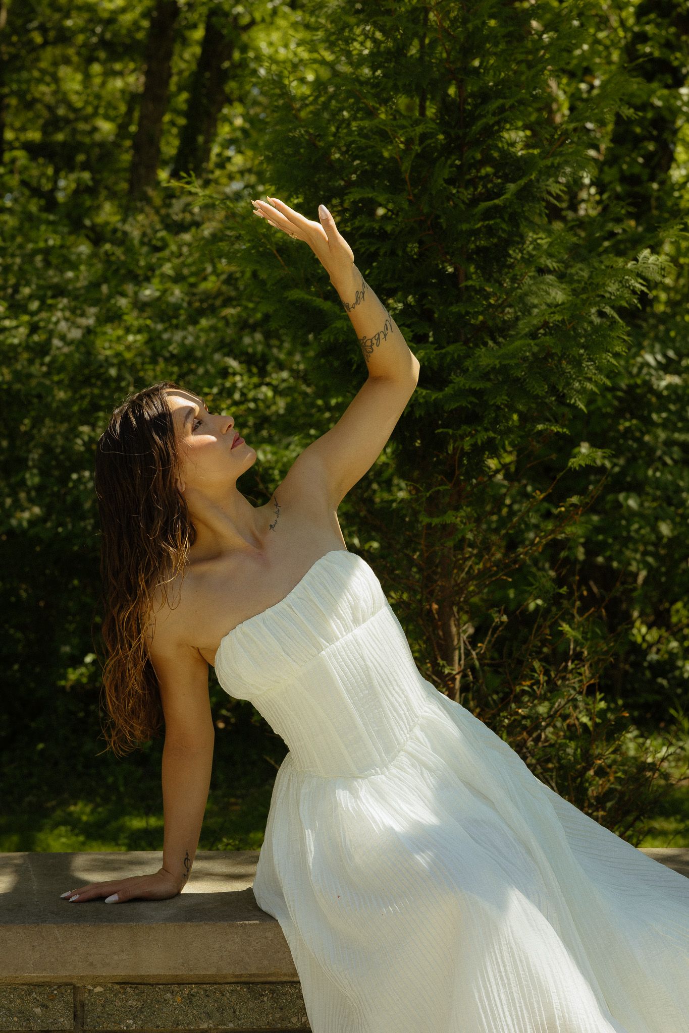 Woman in white strapless dress poses gracefully outdoors with arm raised, surrounded by lush green trees in sunlight.