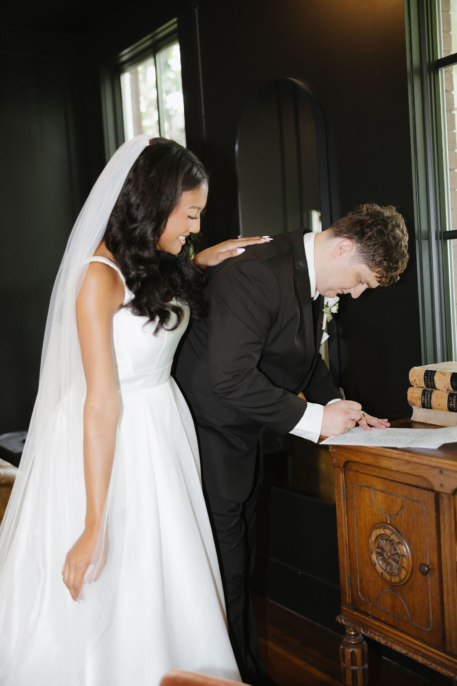 Bride in white gown and veil stands beside groom in black suit signing marriage certificate at wooden desk.