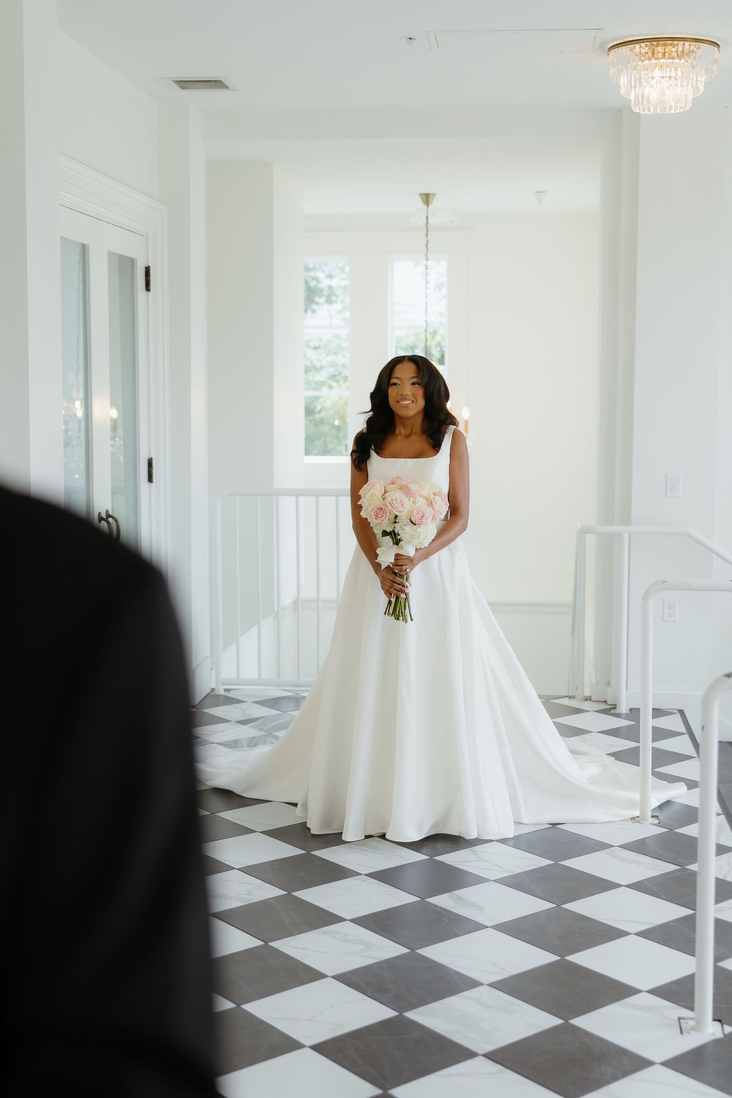 Elegant bride in white ball gown holding pink bouquet stands on checkered floor in bright white venue.