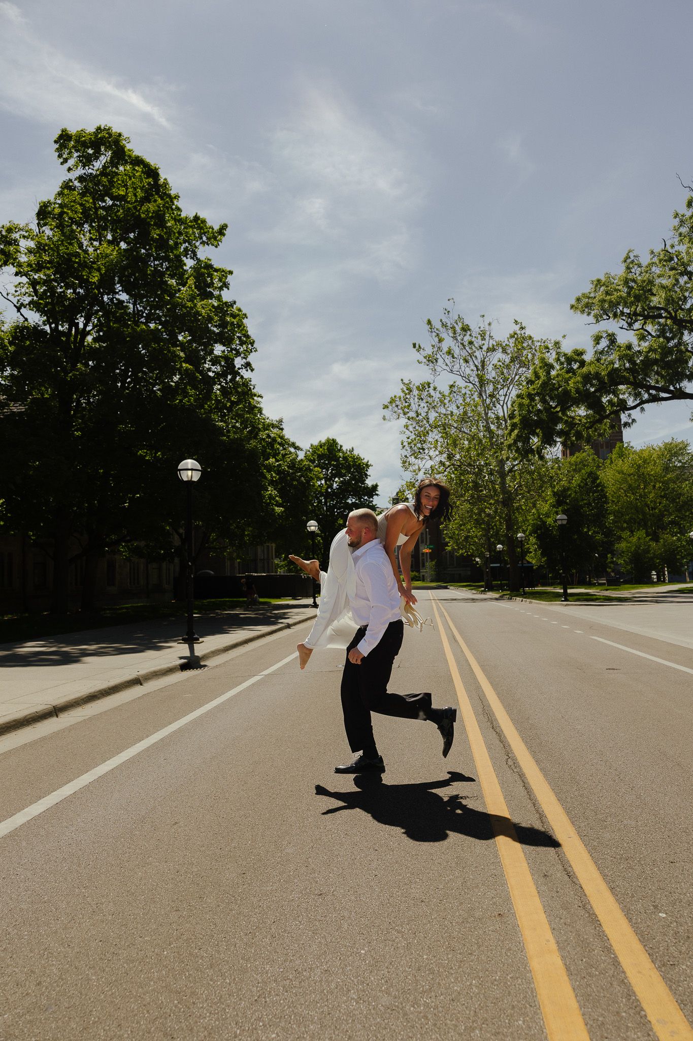A joyful couple in formal attire playfully poses on an empty road lined with green trees on a sunny day.