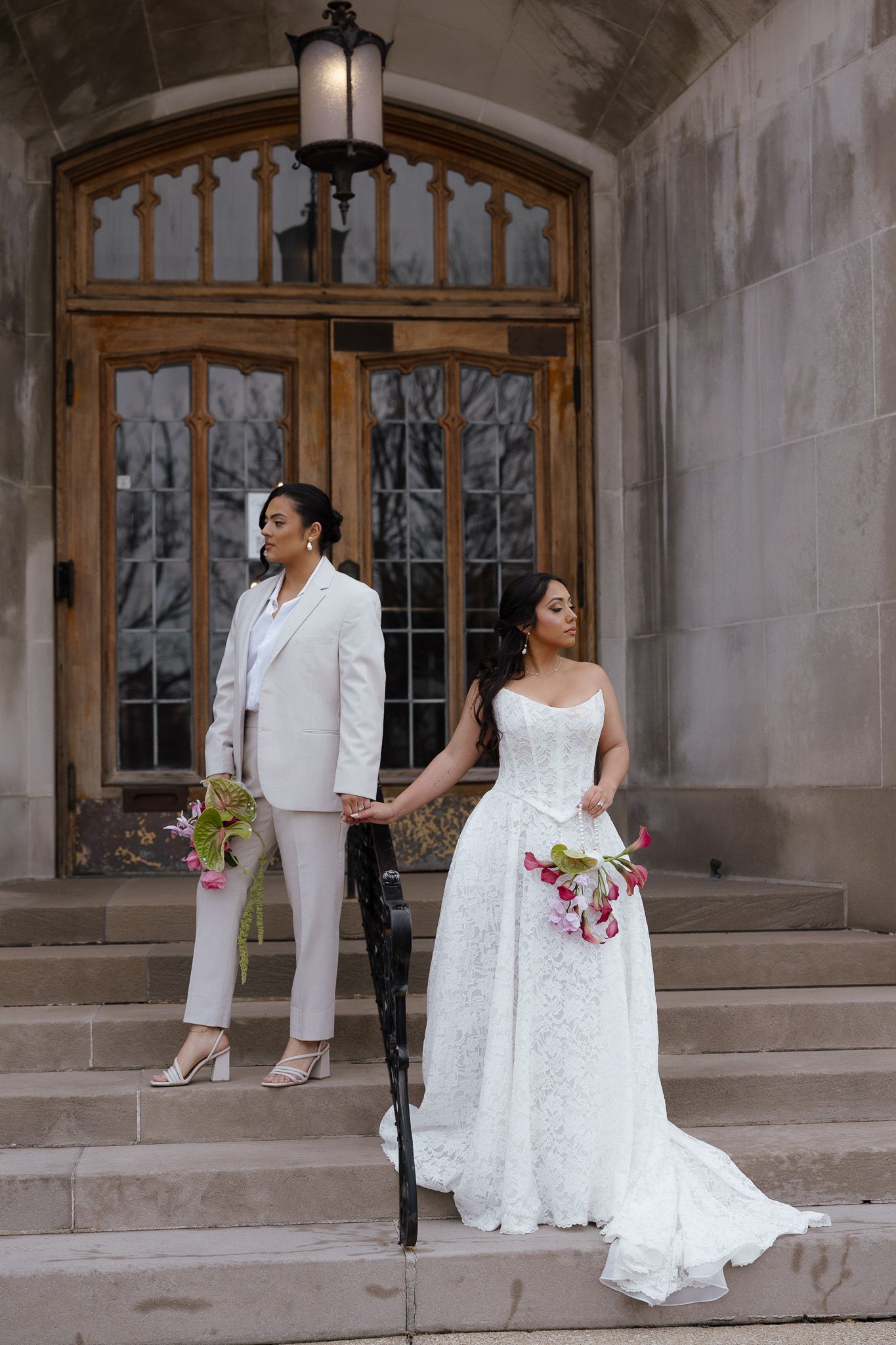 A couple in elegant white wedding attire poses together on stone steps near arched windows.