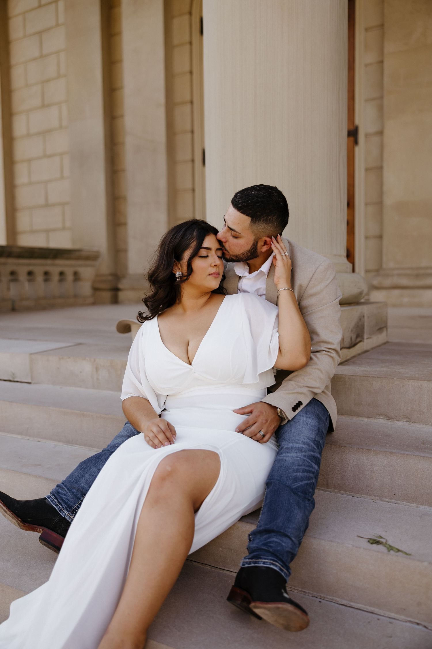 Romantic couple in white clothing poses intimately under stone columns.