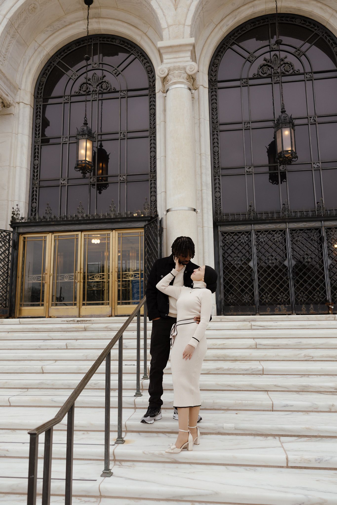 Couple embracing on marble steps of elegant building with arched windows and ornate lanterns.