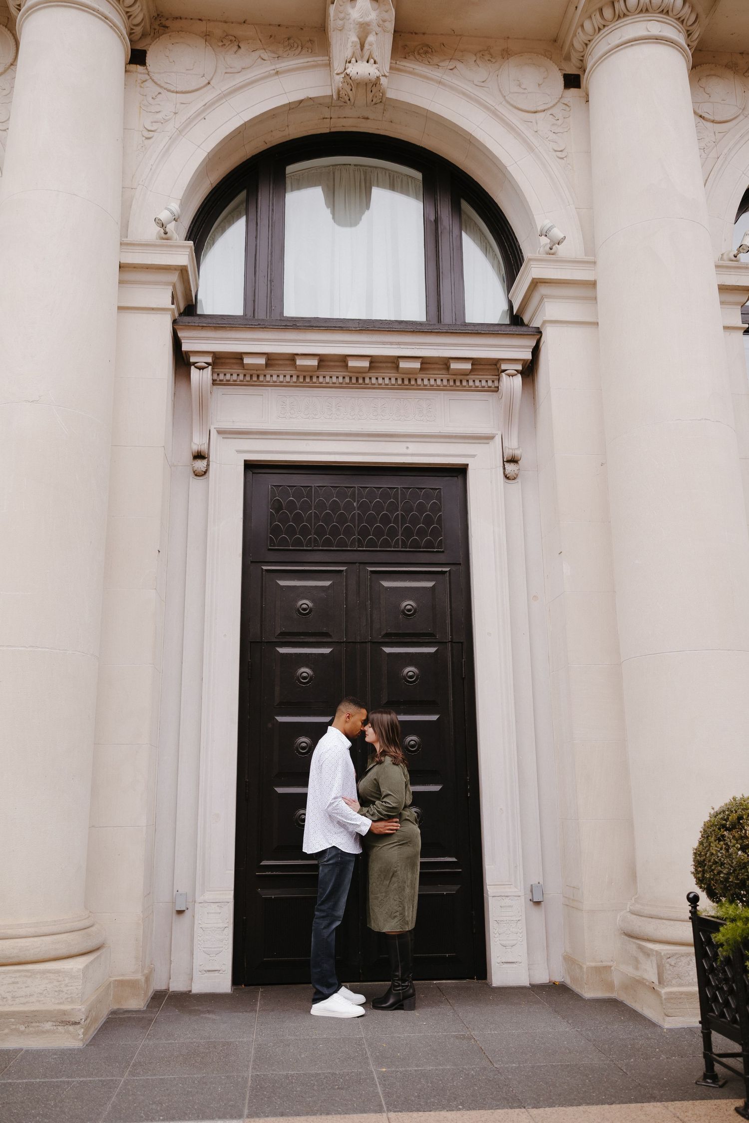 A couple poses romantically by ornate black doors of a classical building.