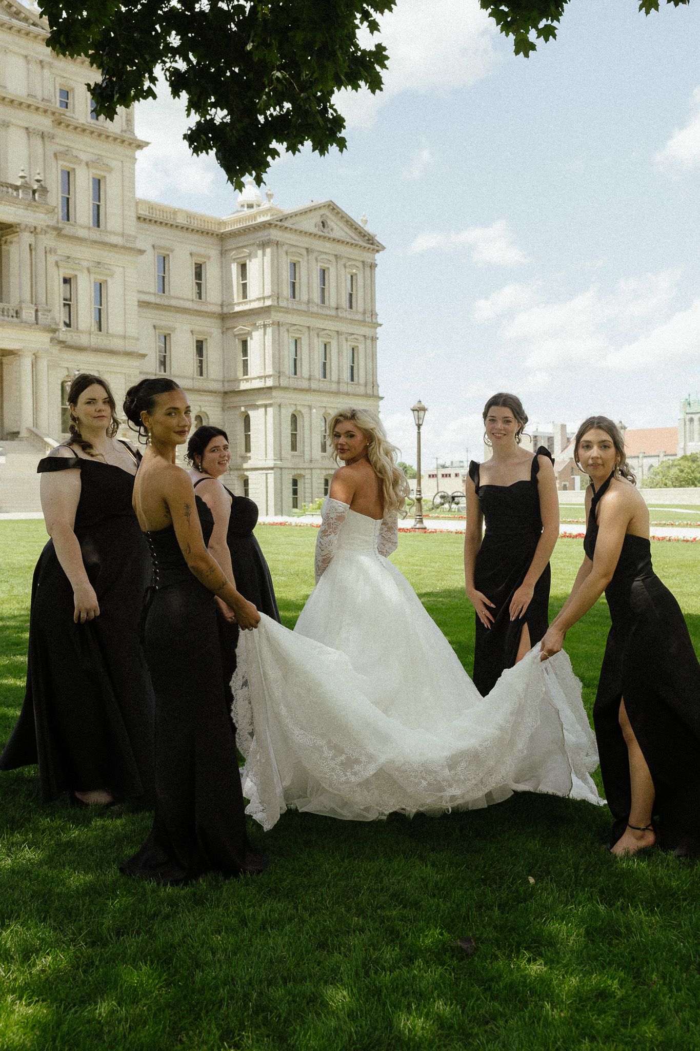 Bride in white gown with bridesmaids in black dresses holding her train outside a grand building.