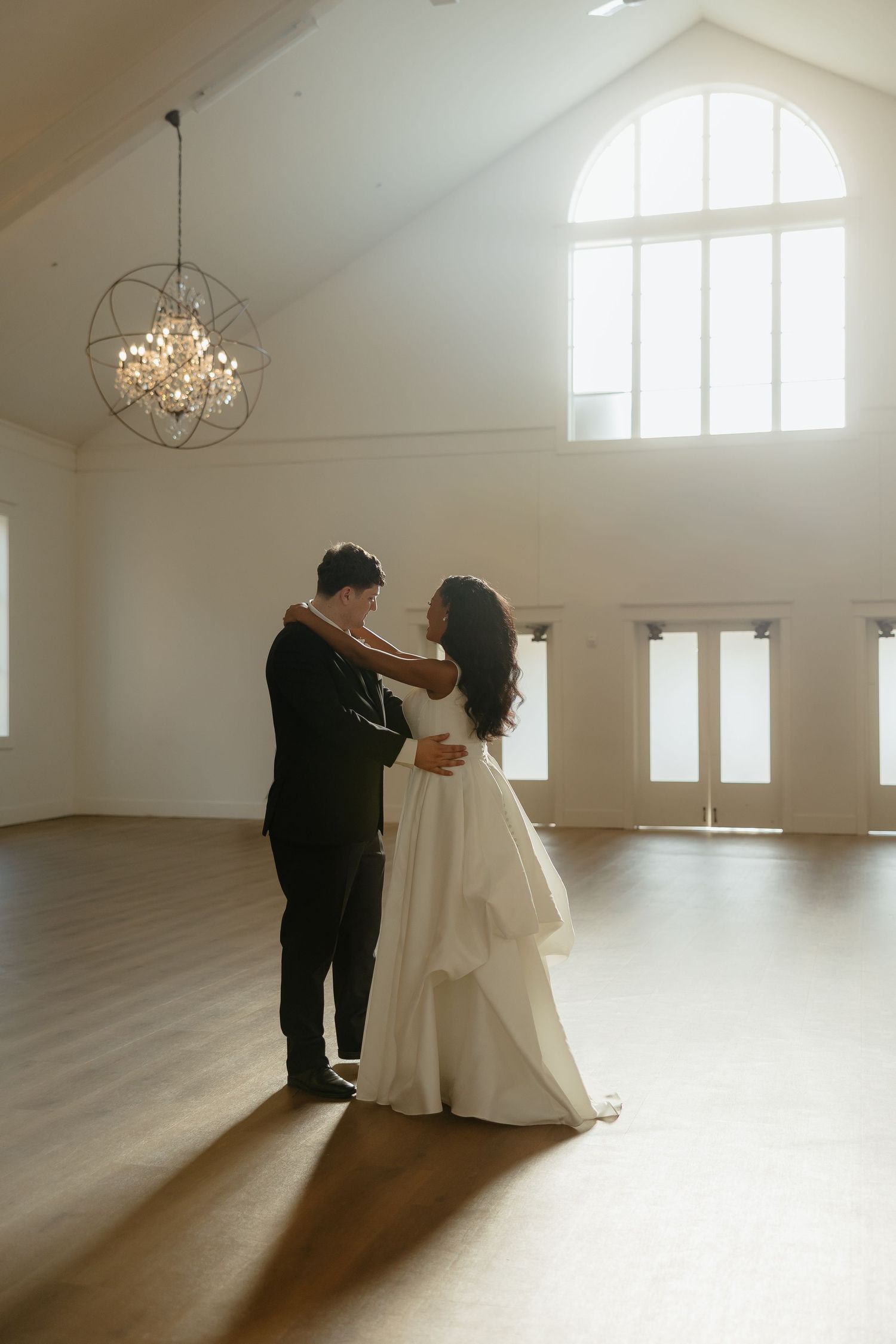 Bride and groom share first dance in elegant white venue with hardwood floors and geometric chandelier.