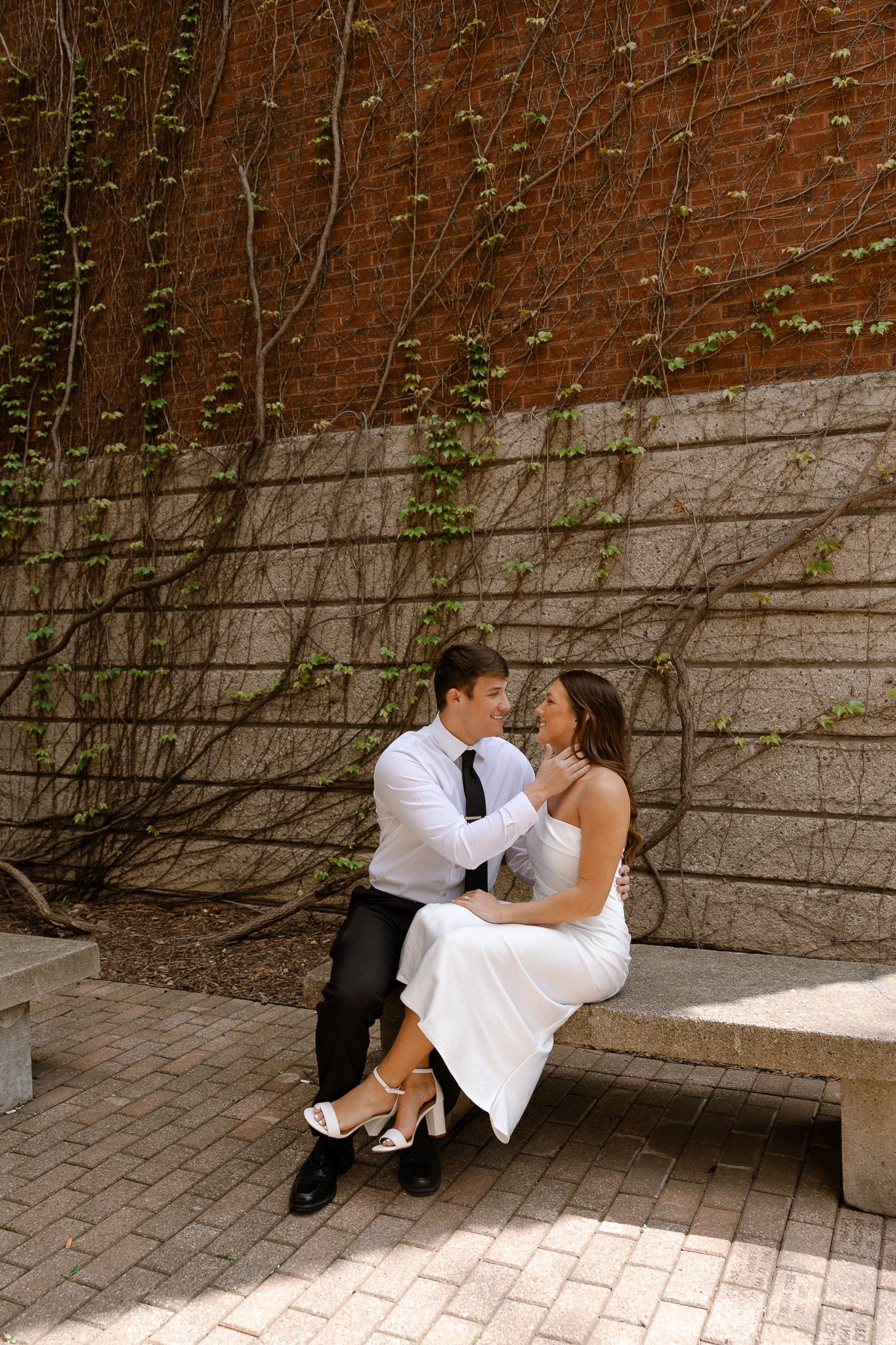 Bride and groom sitting on a stone bench embracing in front of a brick wall with climbing vines.