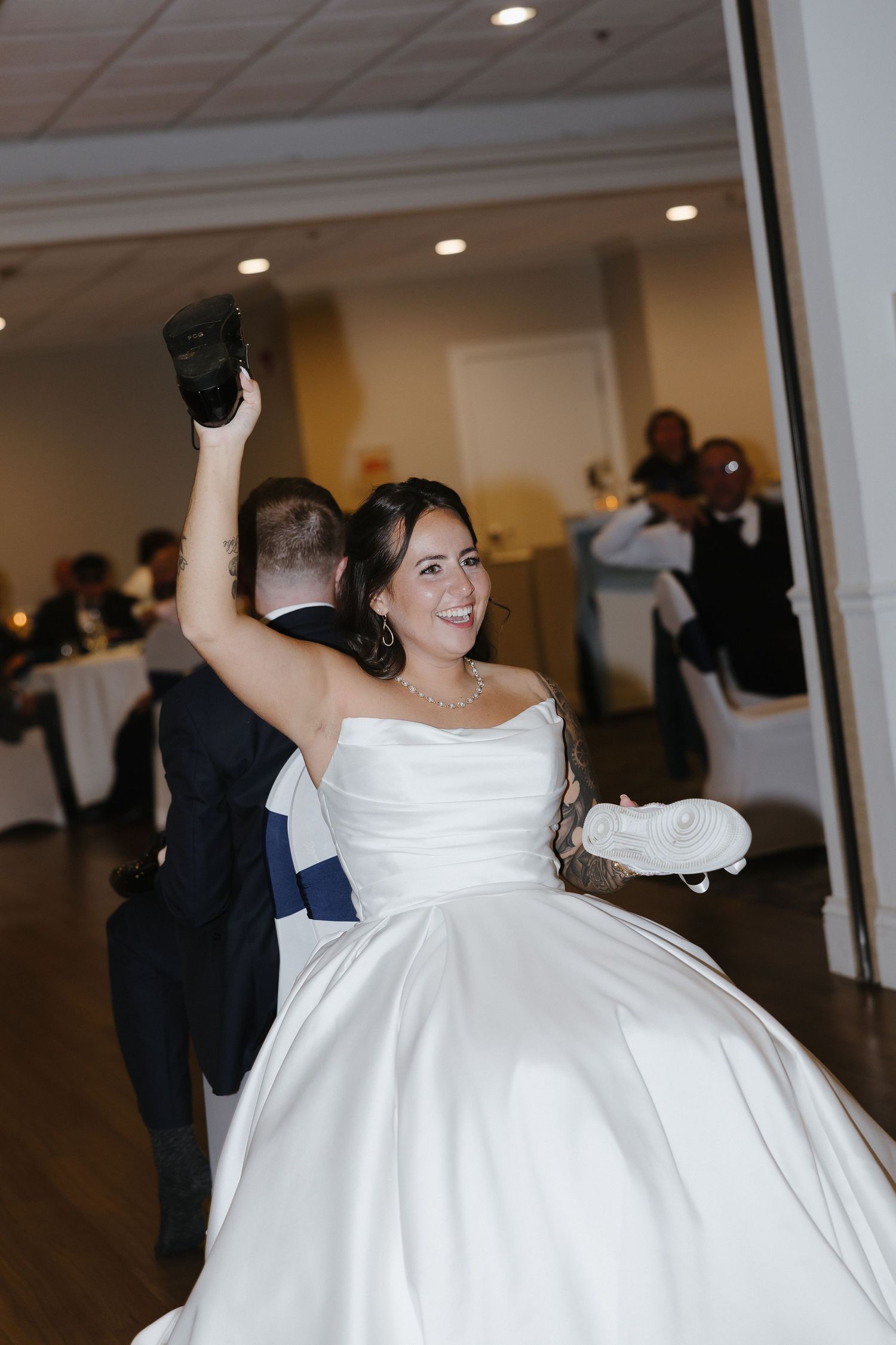 Bride in white ball gown raises shoe triumphantly while entering wedding reception hall, smiling joyfully.